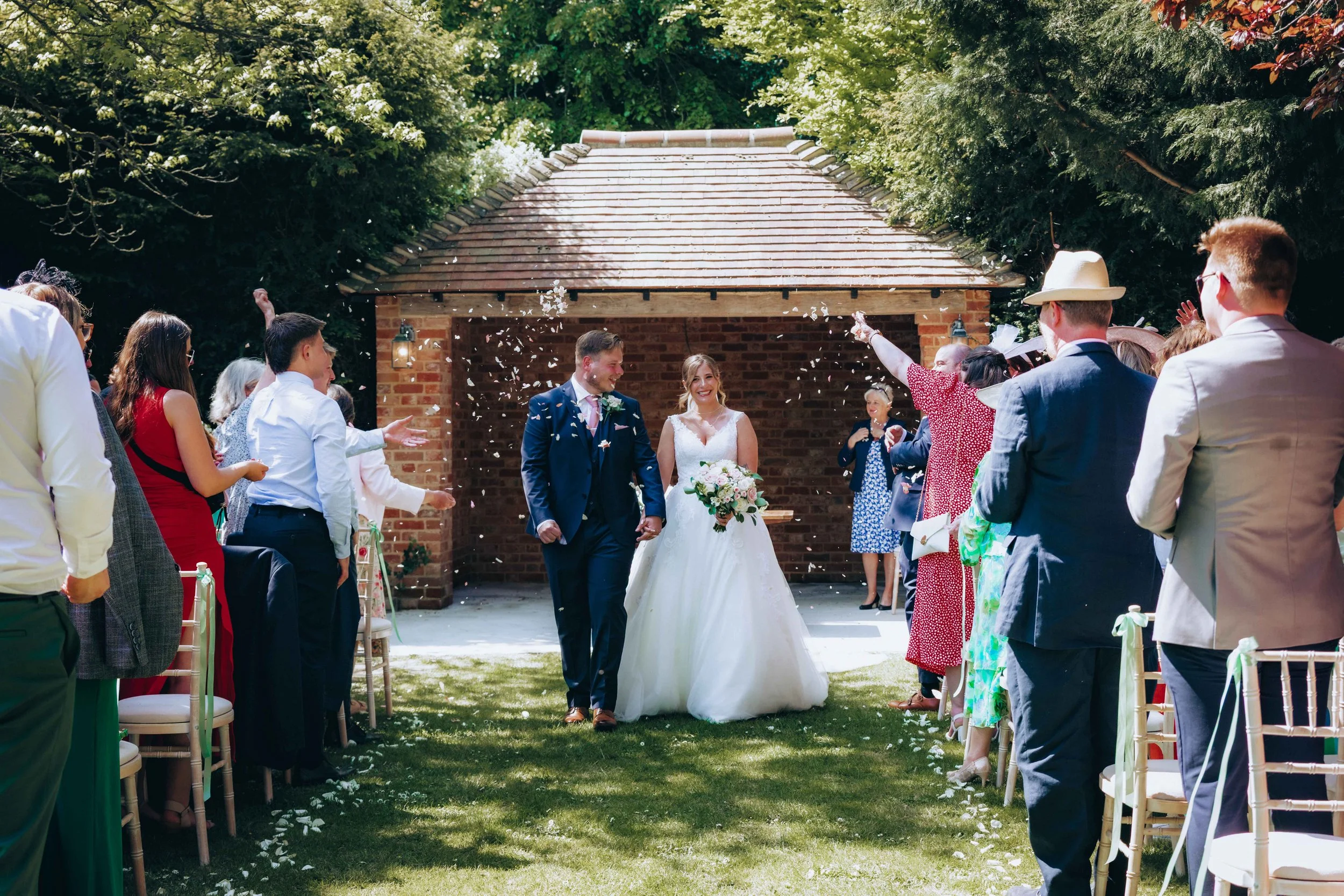 A bride and groom walking hand in hand down the aisle at an outdoor wedding ceremony. Guests are standing on both sides, celebrating and tossing flower petals. The setting includes a brick structure and lush green trees.