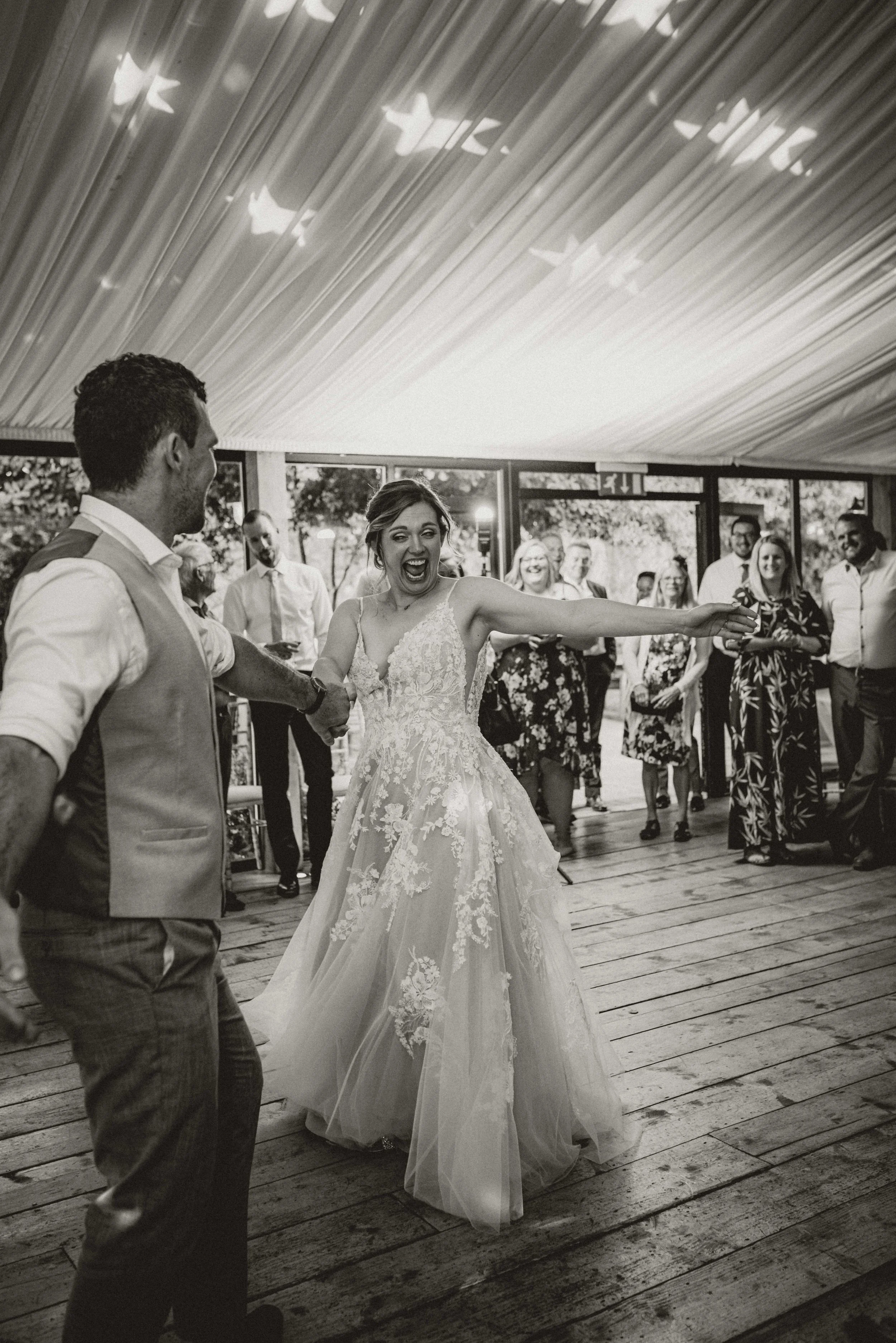 A bride and groom dancing at a wedding reception. The bride is wearing a lace wedding gown and smiling, while the groom is dressed in a vest and shirt. Guests are watching and smiling in the background inside a decorated venue.