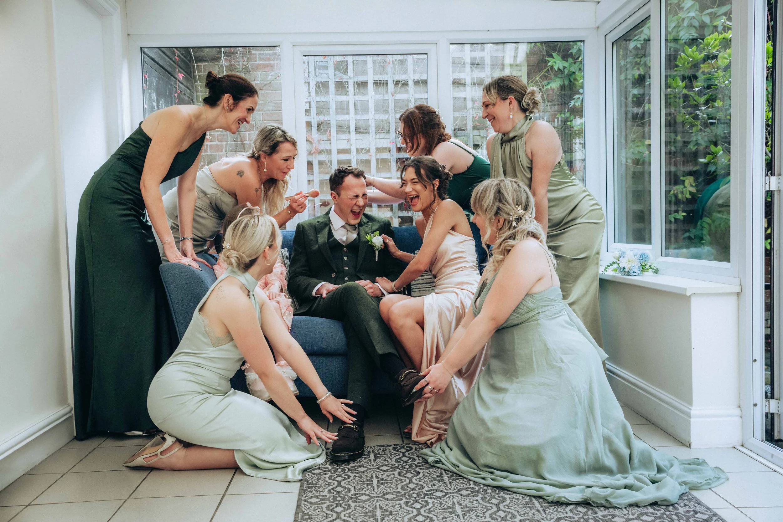 A group of women in elegant dresses and a man in a suit gather in a conservatory or sunroom, smiling and laughing as they help the groom put on his shoe for a wedding or special occasion.