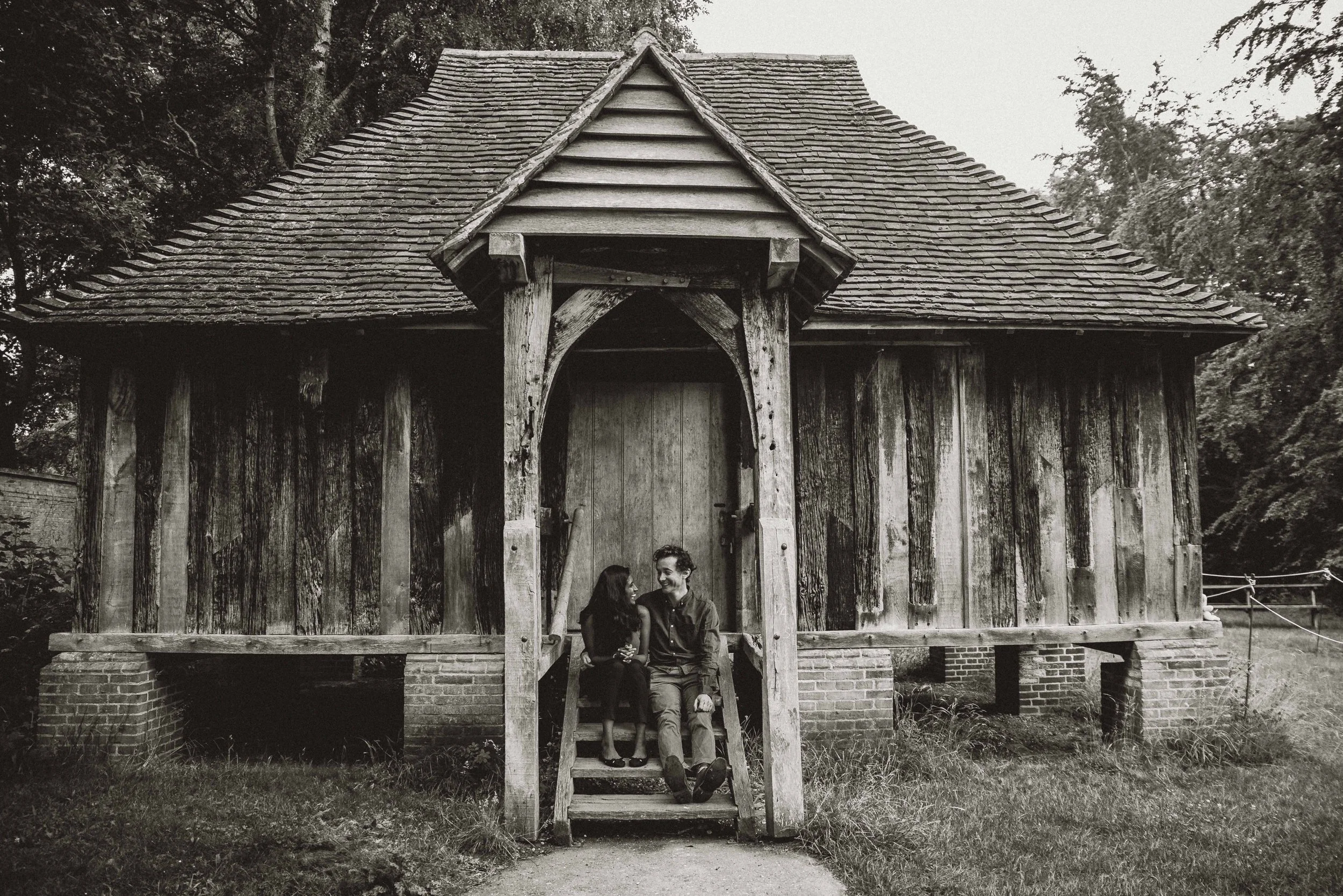 A couple looking at each other smiling while they are sitting on the steps in front of an old wooden house near Cambridge