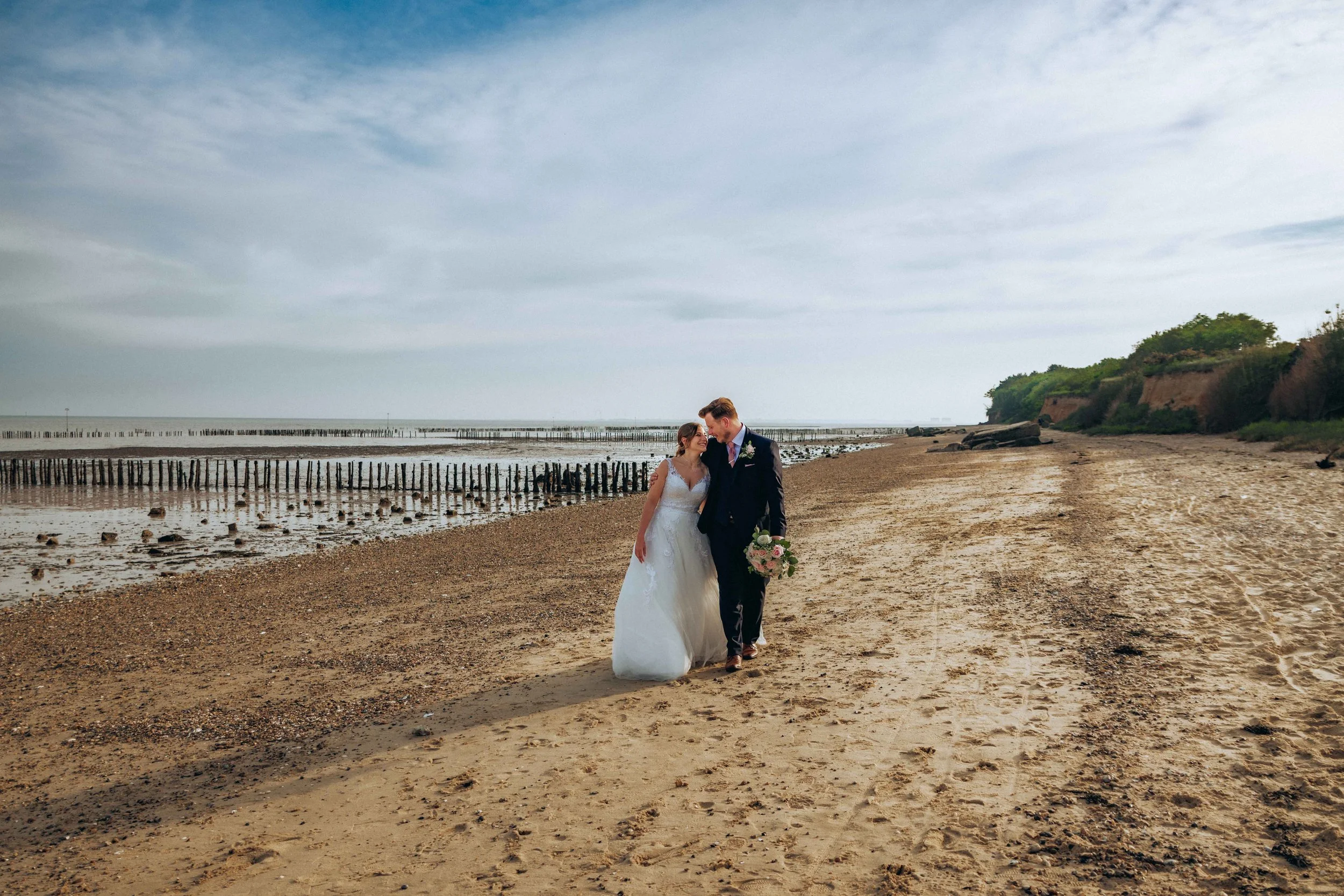 A bride and groom walking together on a sandy beach, with the bride holding a bouquet of flowers and both dressed in wedding attire, near the shoreline with distant cliffs and cloudy sky.