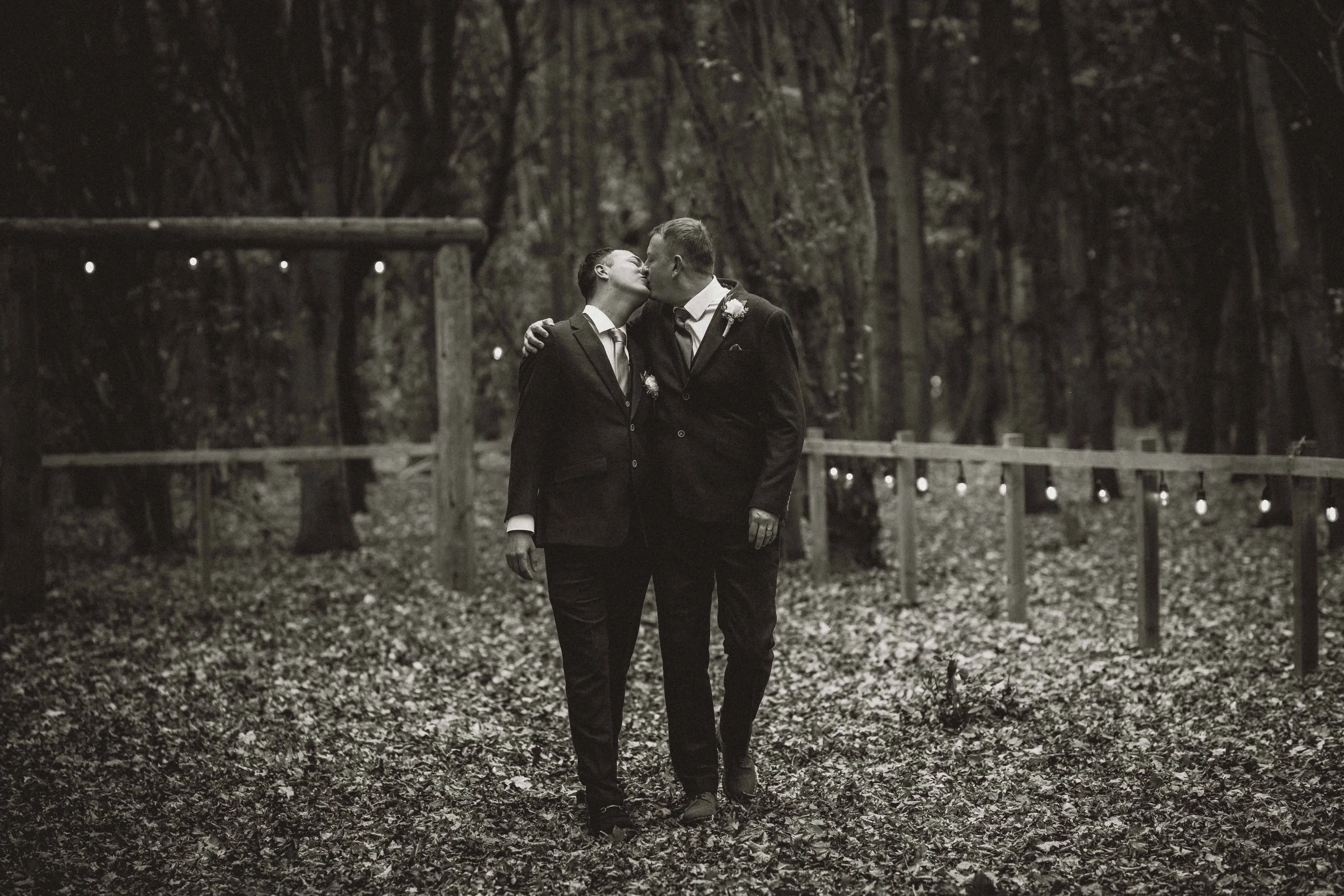 Two men in suits sharing a kiss in a wooded outdoor setting, walking along a trail surrounded by trees and fallen leaves at dusk or night.