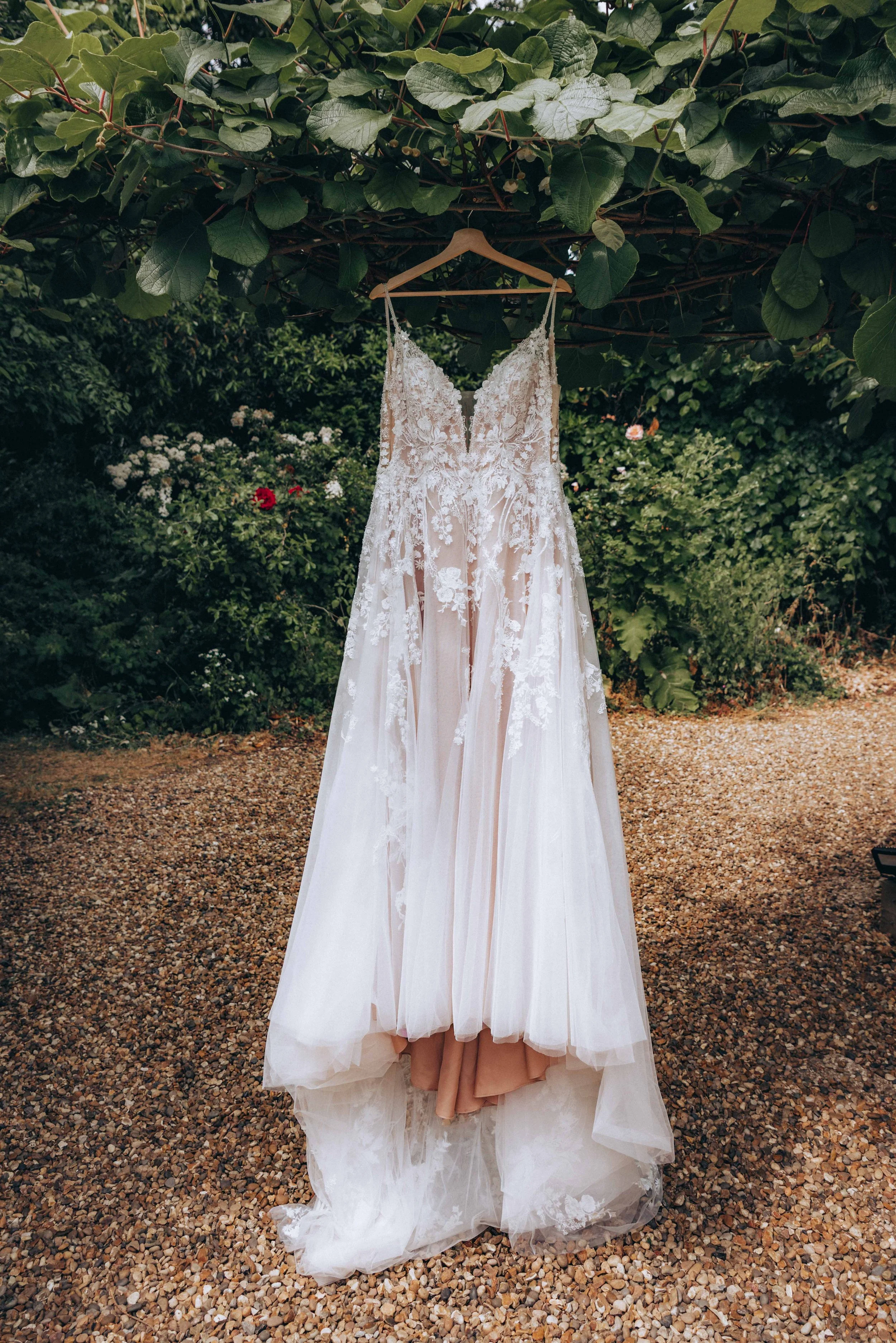 Back view of a wedding dress hanging on a hanger under a leafy tree in an outdoor garden setting.
