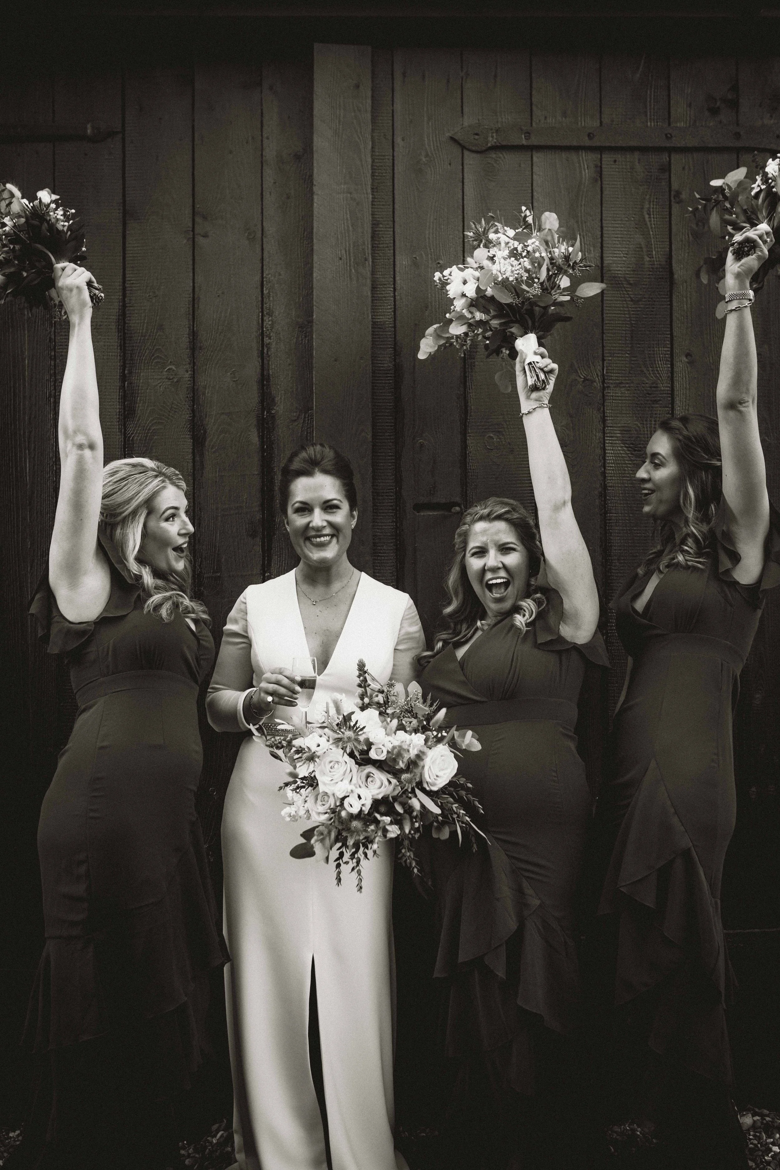 Four women, with three holding bouquets of flowers and one holding a drink, standing in front of a wooden wall, celebrating at a wedding.