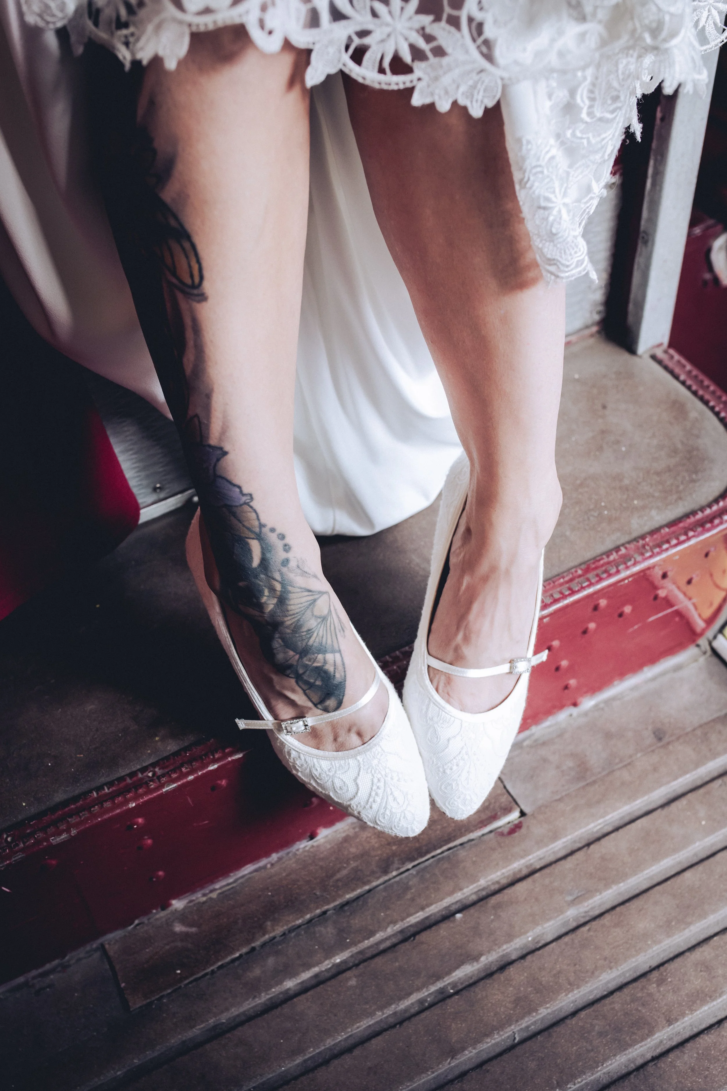Person wearing white lace wedding shoes with a floral lace dress and tattooed leg.
