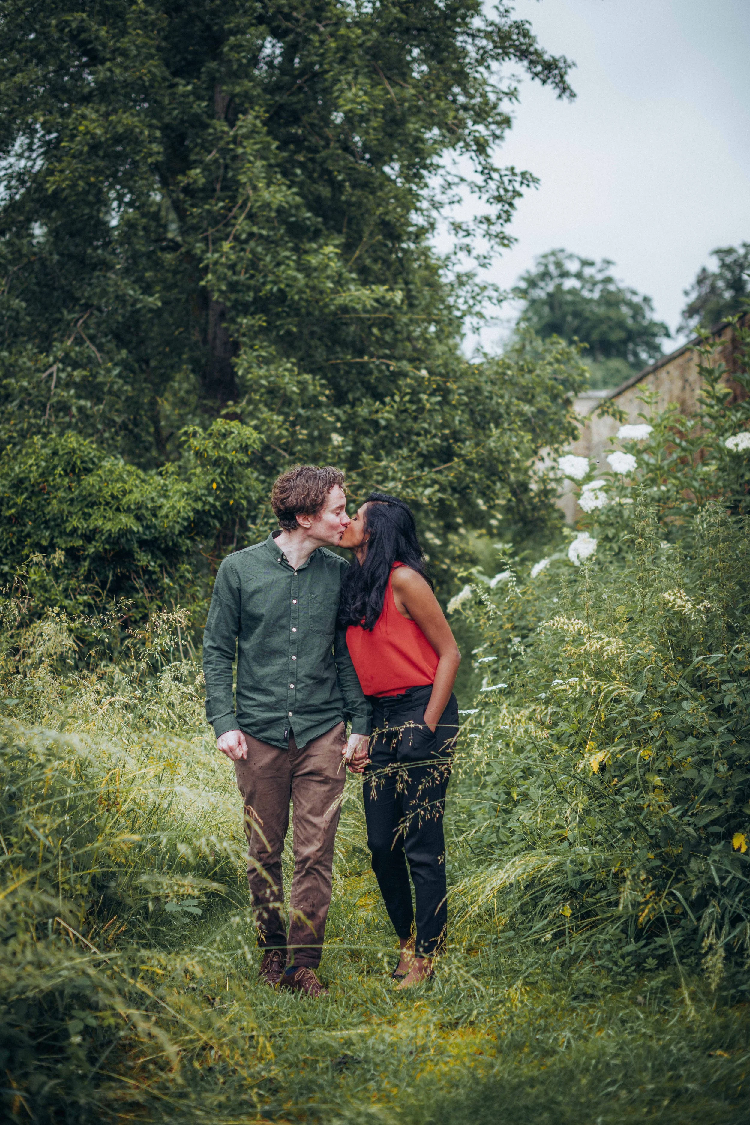 A couple leaning in for a kiss while holding hands surrounded by lush greenery in a park near Cambridge during an engagement session