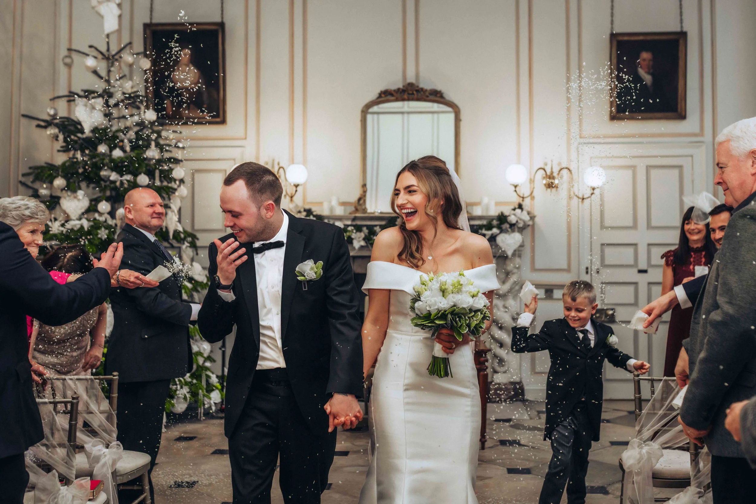 Bride and groom holding hands, walking through a celebration with confetti at Gosfield Hall in Halstead, surrounded by family and friends in a decorated wedding hall.