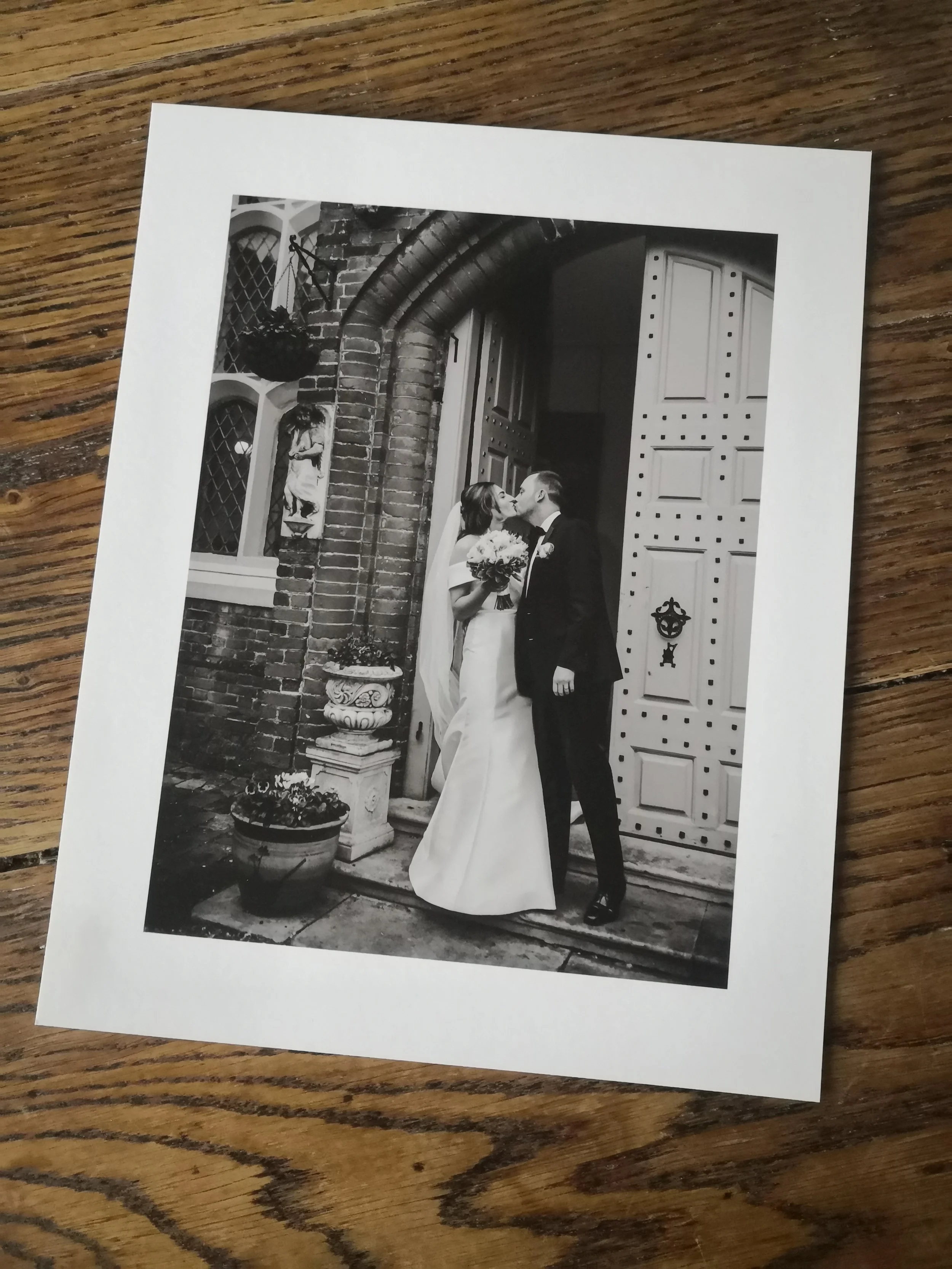 A black and white photograph of a bride and groom sharing a kiss outside a church, with a brick wall, a statue, and decorative flowers around them.