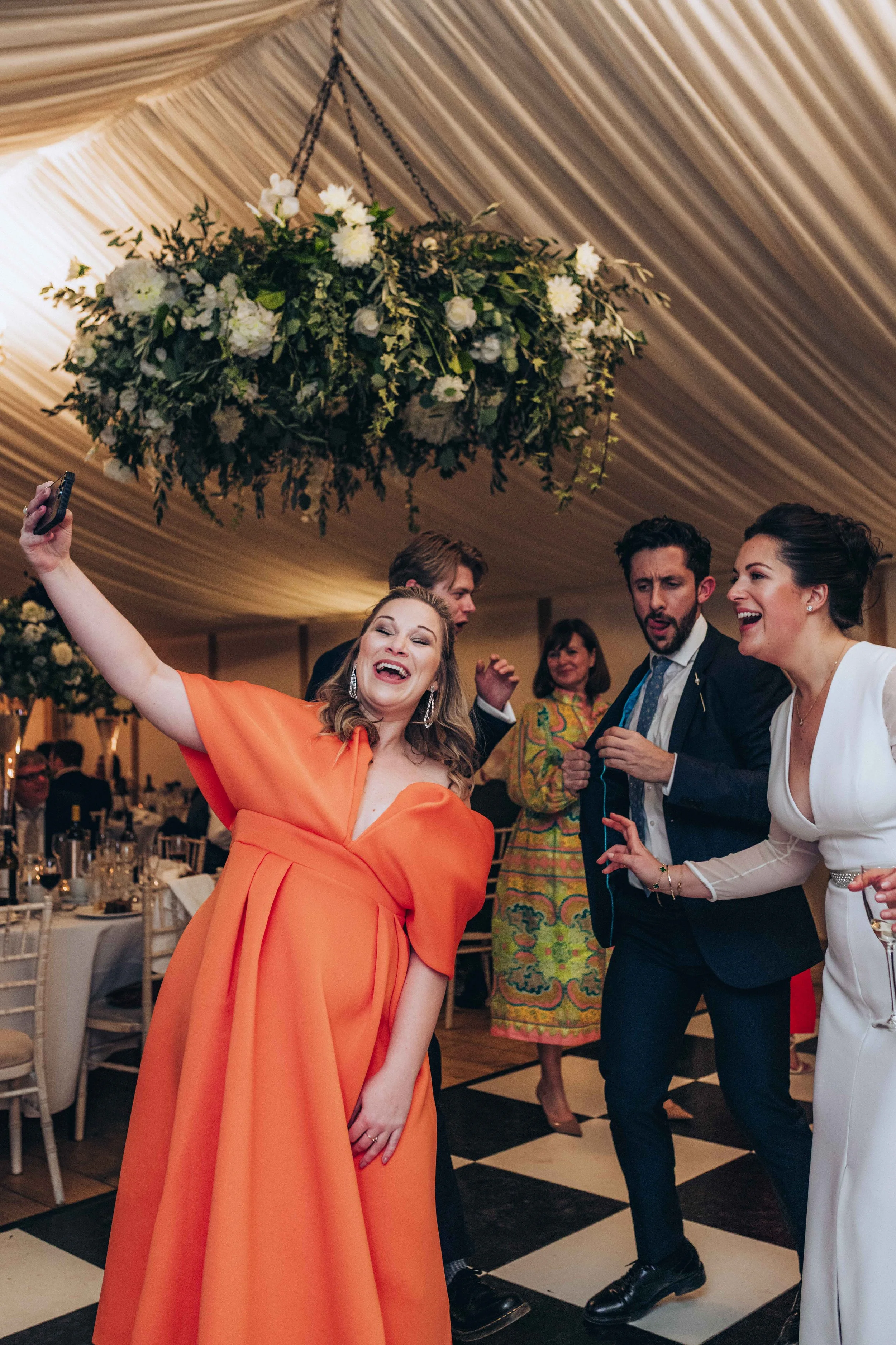 People dancing and celebrating at a wedding reception with a floral chandelier hanging from a draped ceiling.