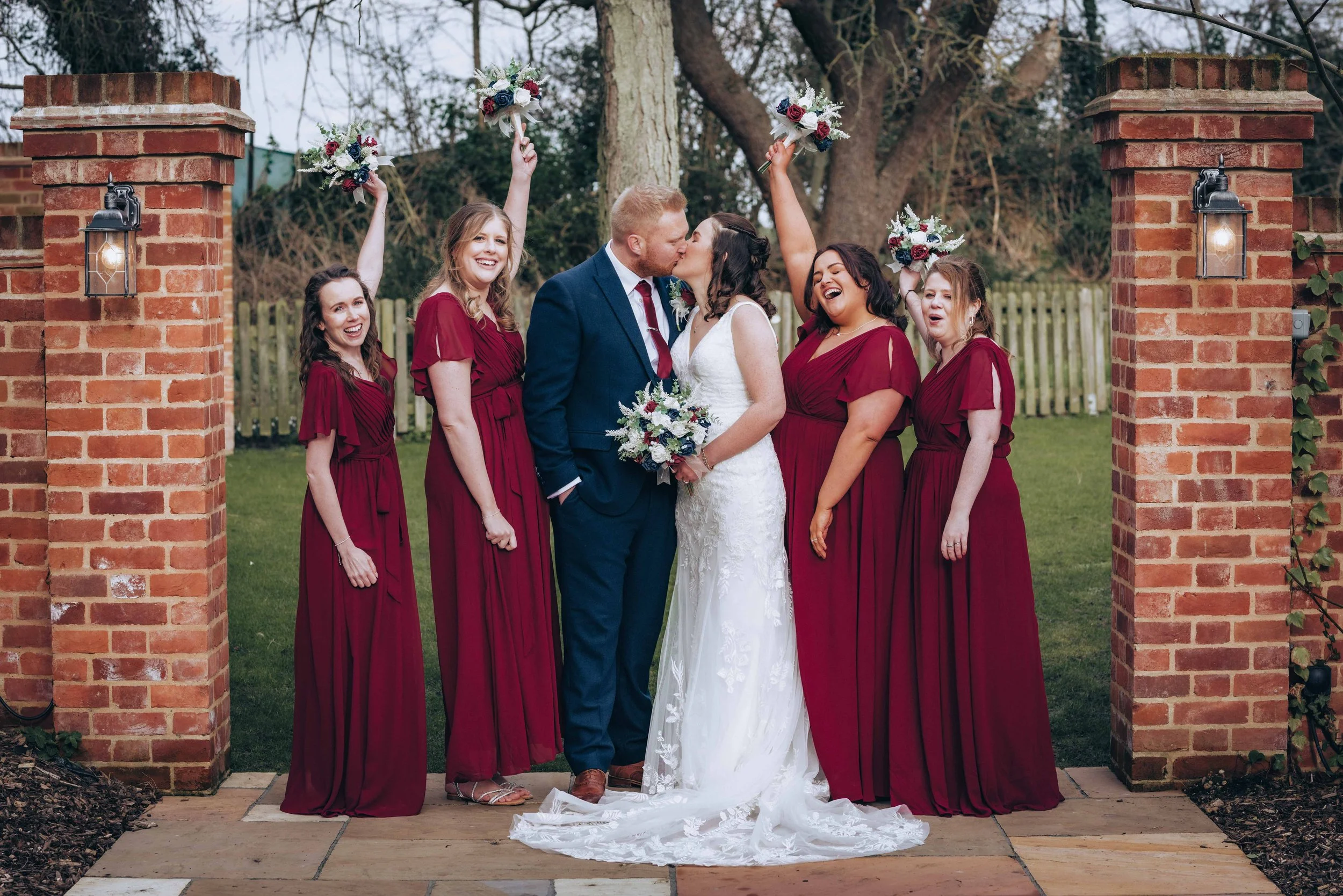A bride and groom kissing during their wedding ceremony, surrounded by five bridesmaids in burgundy dresses, holding bouquets, in an outdoor setting with brick pillars and trees.