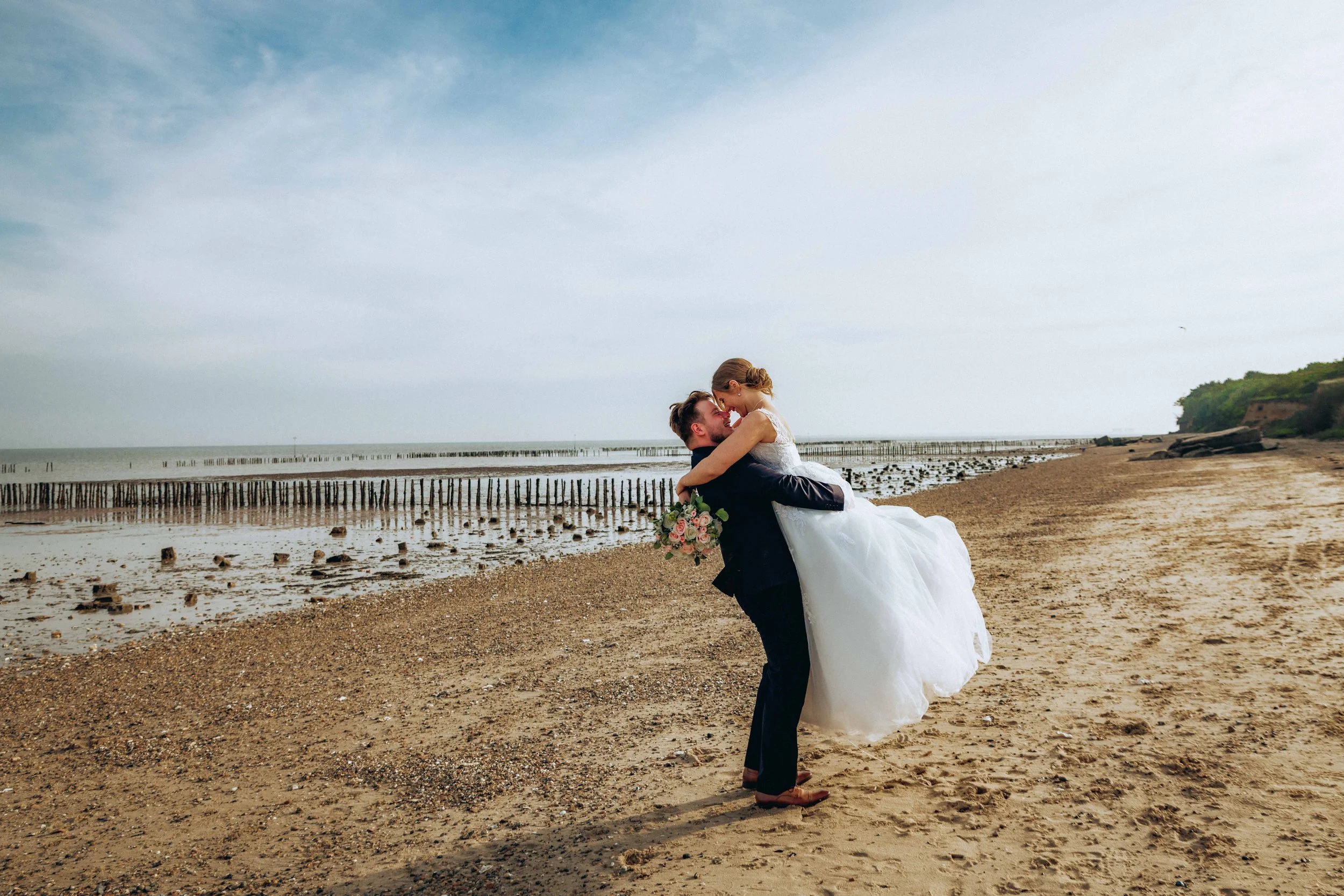 A newlywed couple on the beach, with the man lifting the woman in his arms, both smiling. The woman wears a white wedding dress, holding a bouquet of flowers. The beach has driftwood and wooden posts, with water and a tree-covered hill in the background under a partly cloudy sky.