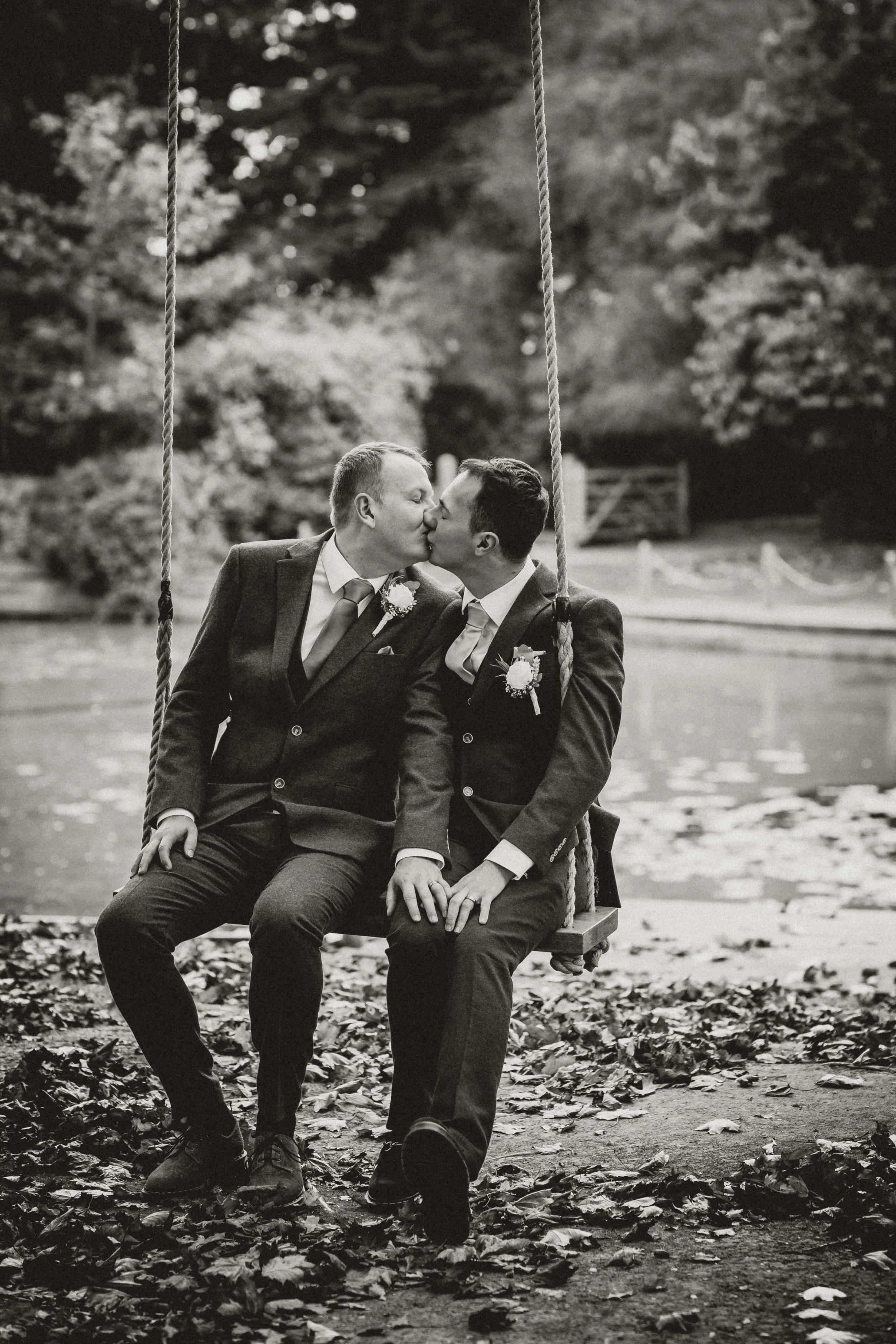 Two men in suits sharing a kiss while sitting on a swing with a backdrop of a park and fall foliage.