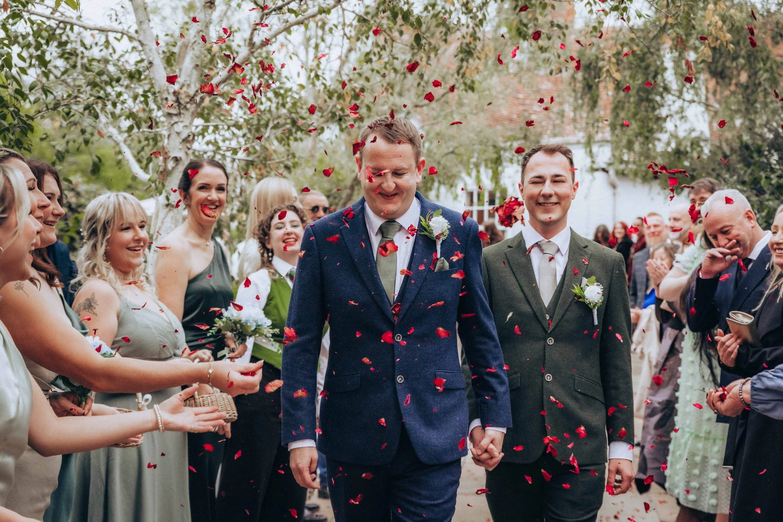 Groom and groom with wedding guests releasing red flower petals outdoors during a wedding celebration.