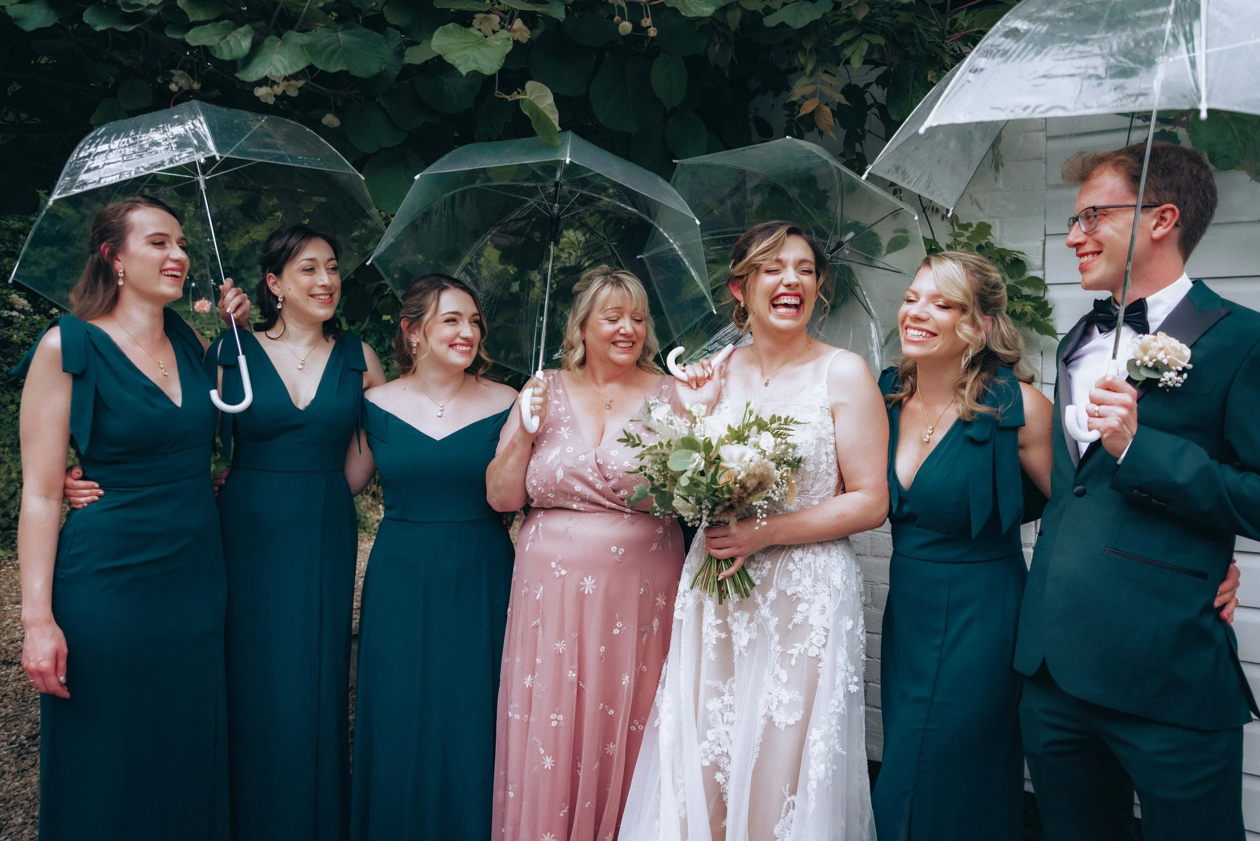 A group of eight people, including women in formal dresses and a man in a tuxedo, standing close together holding transparent umbrellas outdoors on a rainy day. They are smiling and appear to be celebrating a wedding or special event.