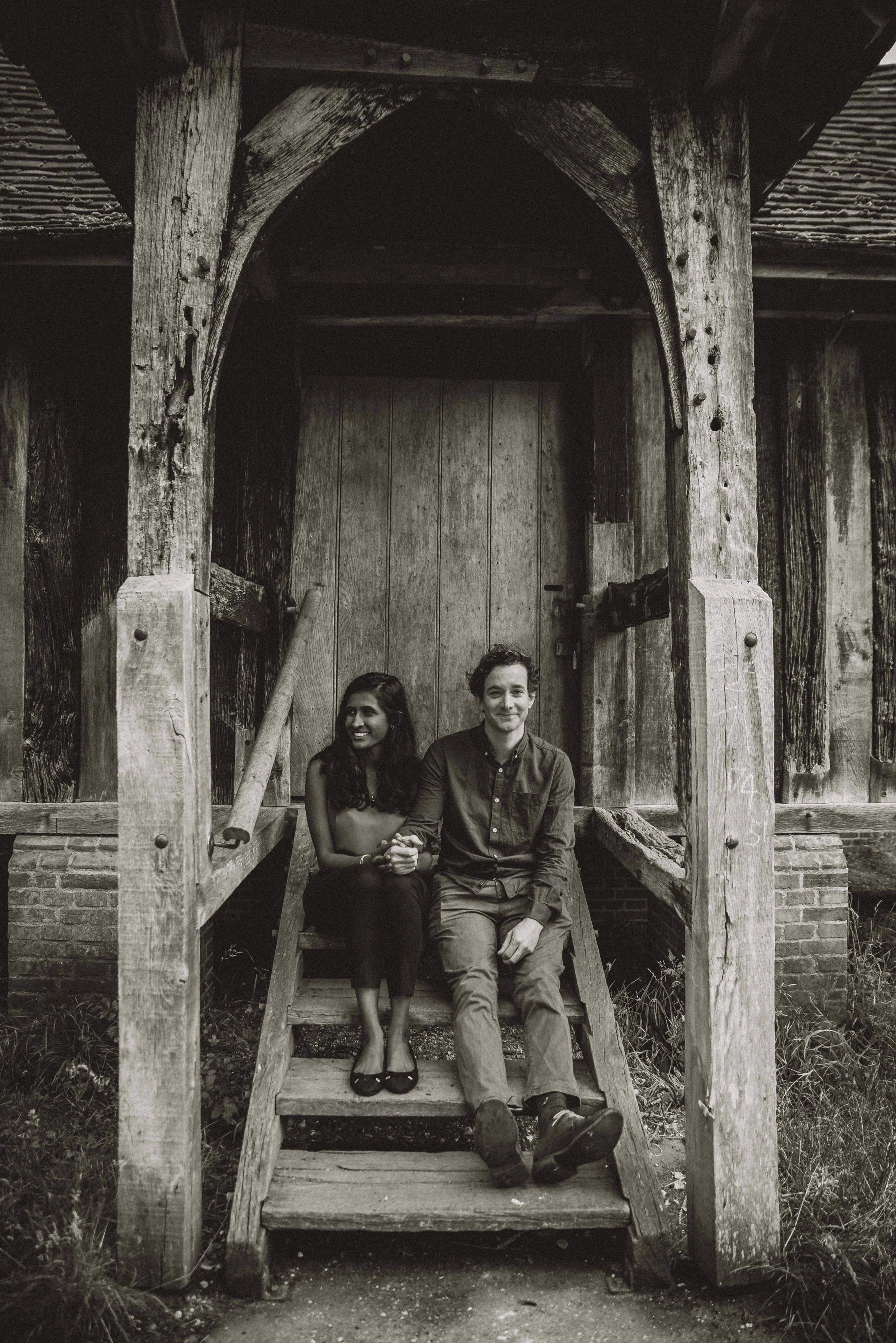 A couple holding hands and sitting down on wooden steps with him looking at the camera in a park near Cambridge during an engagement photo session