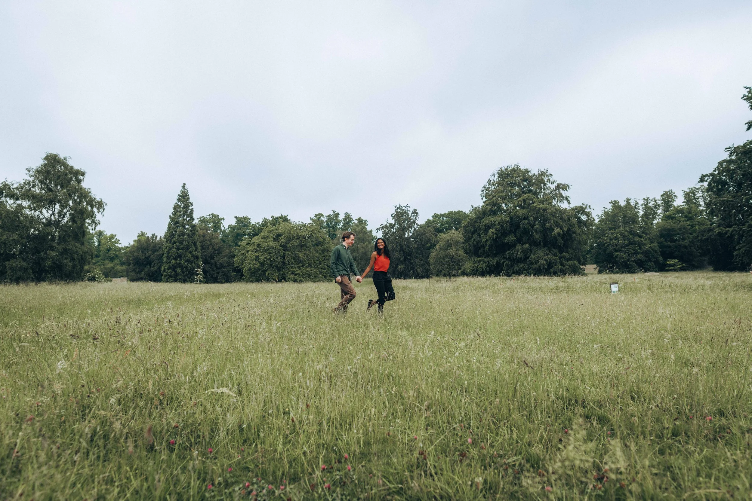 A couple holding hands and walking across a lush meadow with trees in the background near Colchester