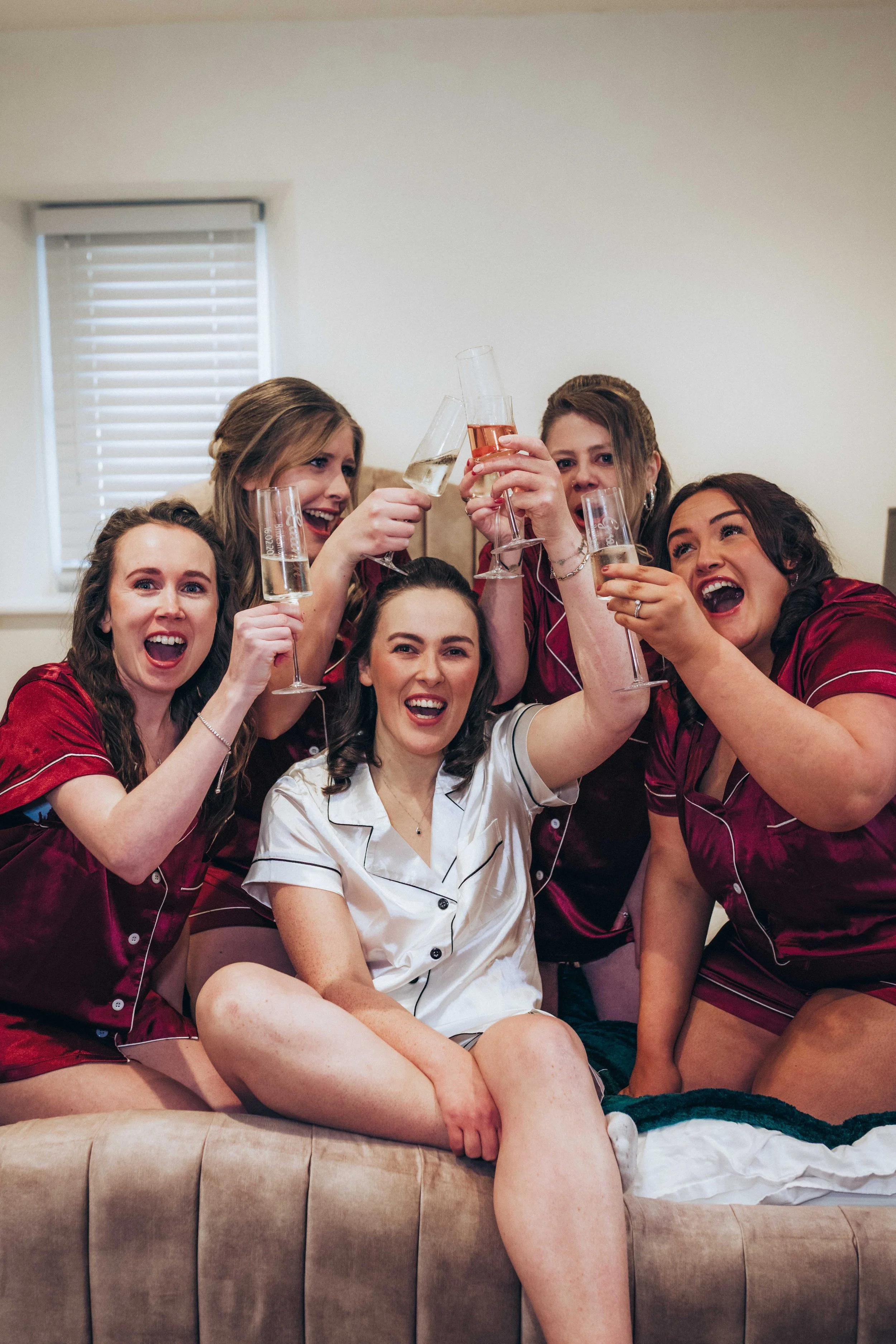 A group of six women in pajamas celebrating and raising glasses of champagne on a beige couch in a living room with a window with blinds in the background.