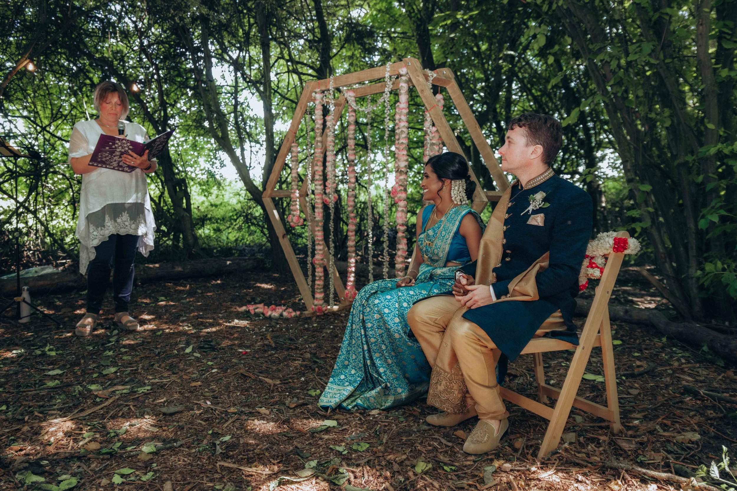 A couple dressed in traditional Indian wedding attire sitting on chairs under a canopy of trees, with a woman reading from a book or script nearby during an outdoor wedding ceremony surrounded by nature.