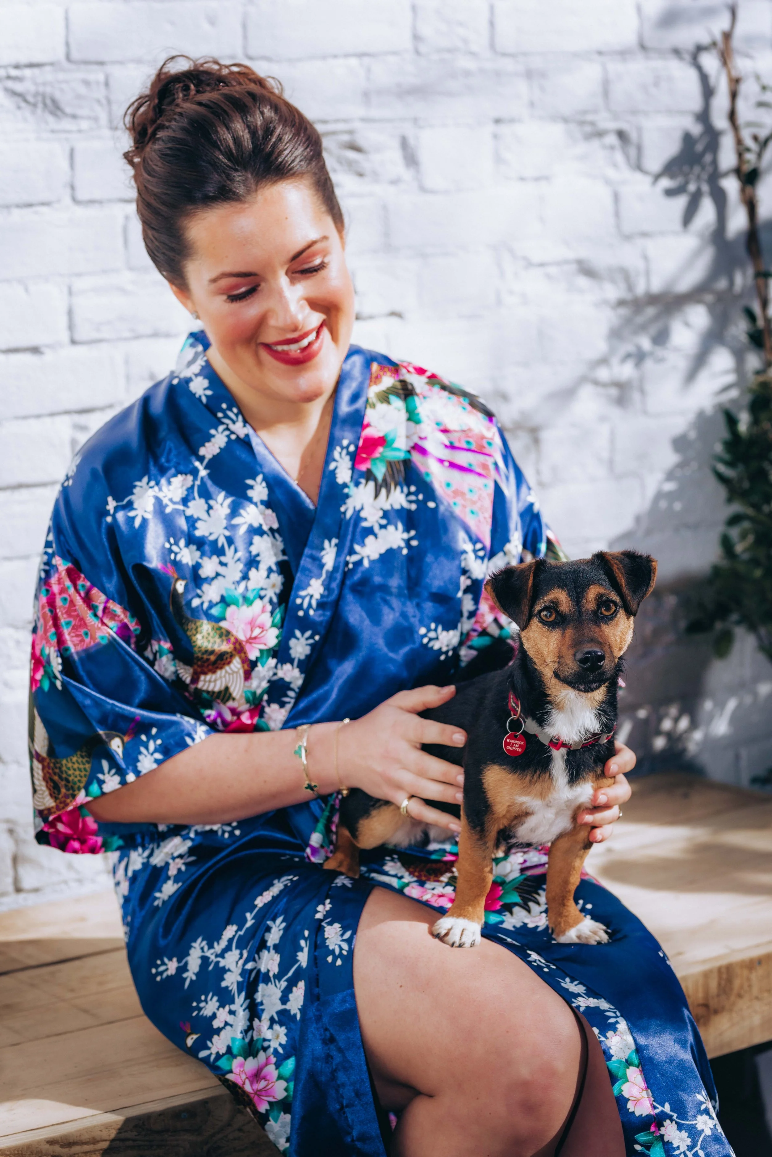 A woman with dark hair styled in an updo, wearing a blue floral kimono, sitting on a wooden bench, holding a small black and brown dog with a red collar, against a white brick wall.
