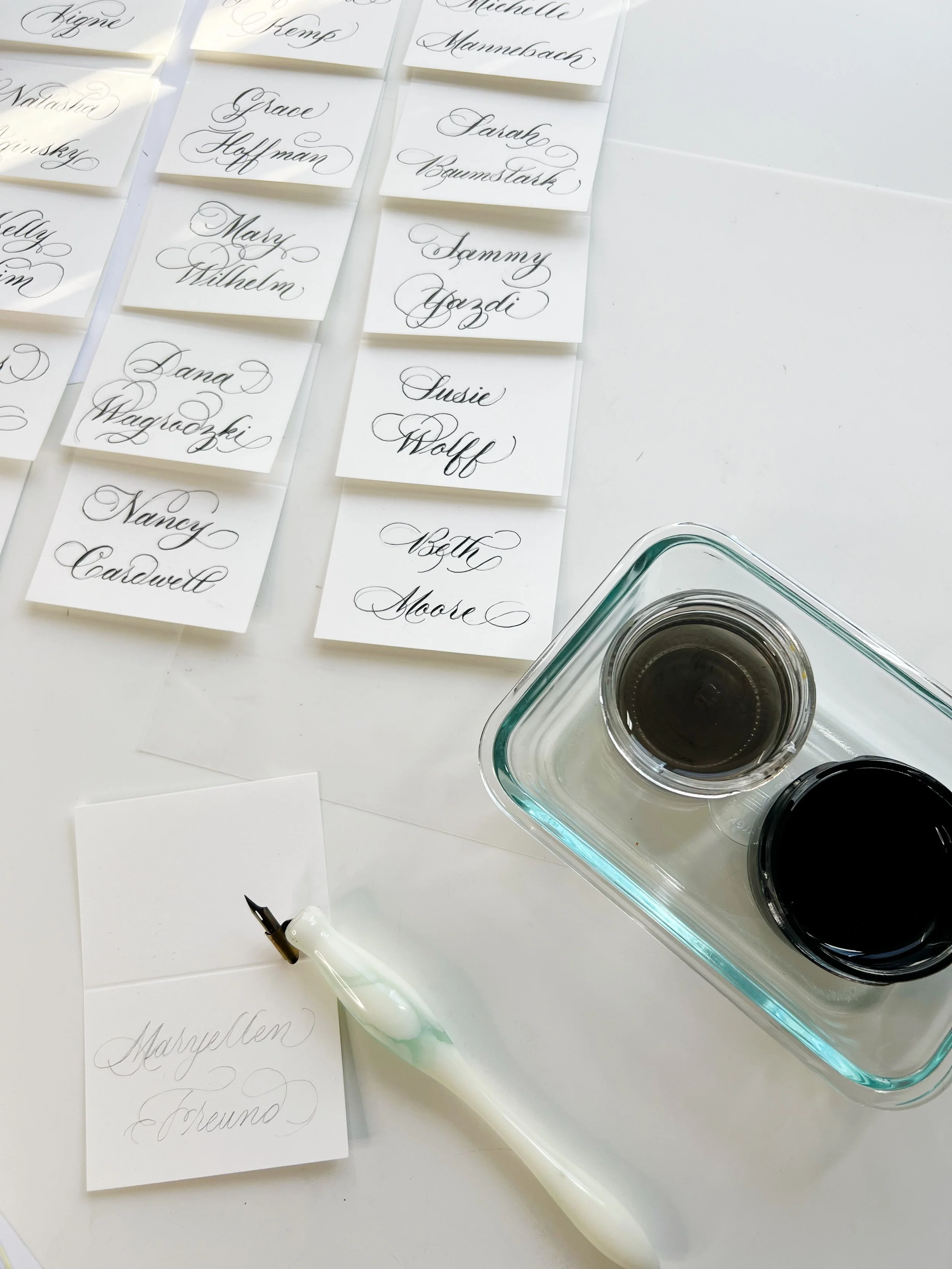 Calligraphy practice setup with sheets of paper featuring names written in elegant script, a white dip pen with a black nib, and a glass dish holding black ink along with a small container of gray ink on a white surface.