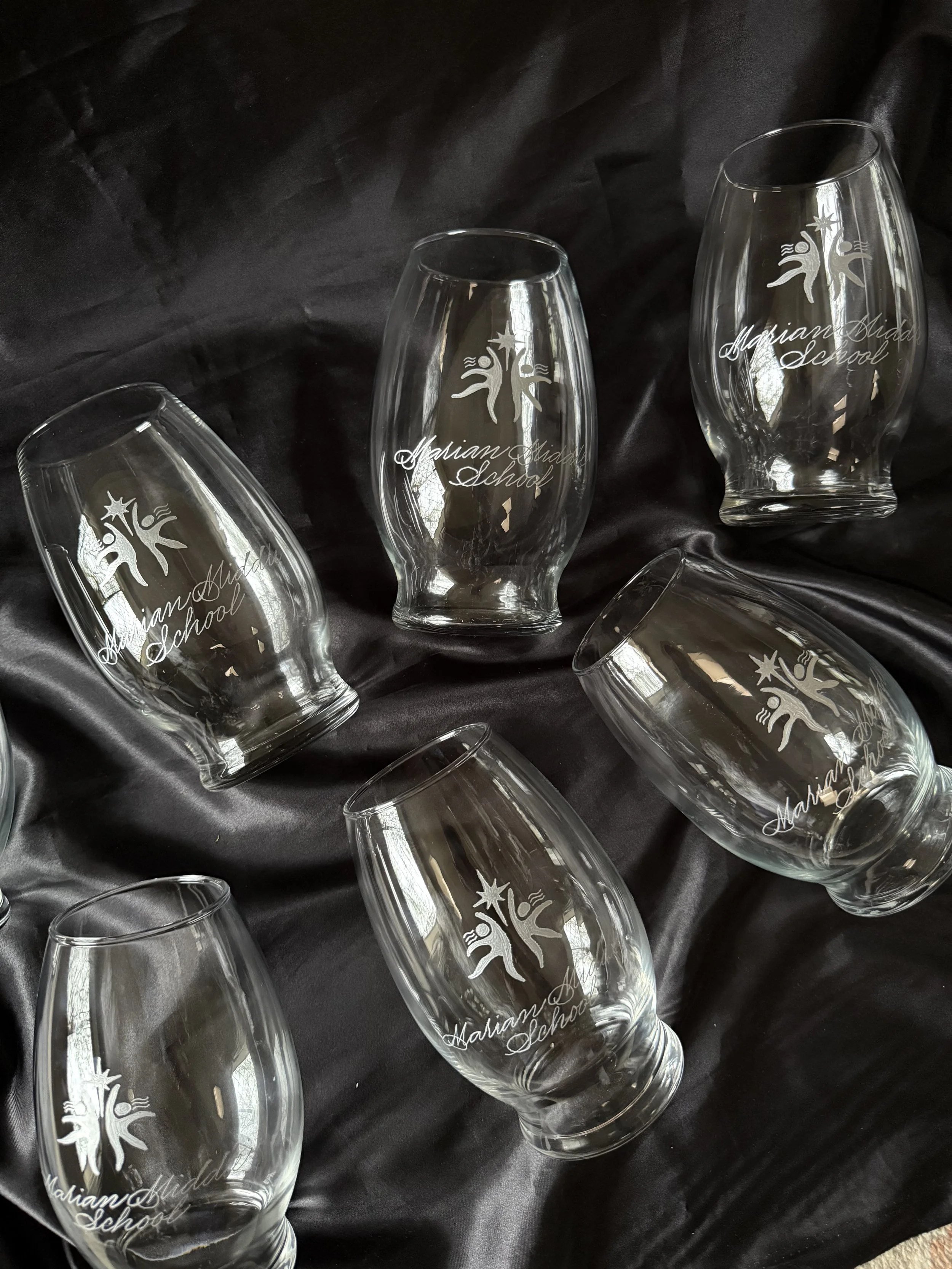 Multiple clear glass floral vases with 'Marian Middle School' hand engraved, arranged on a dark fabric surface.
