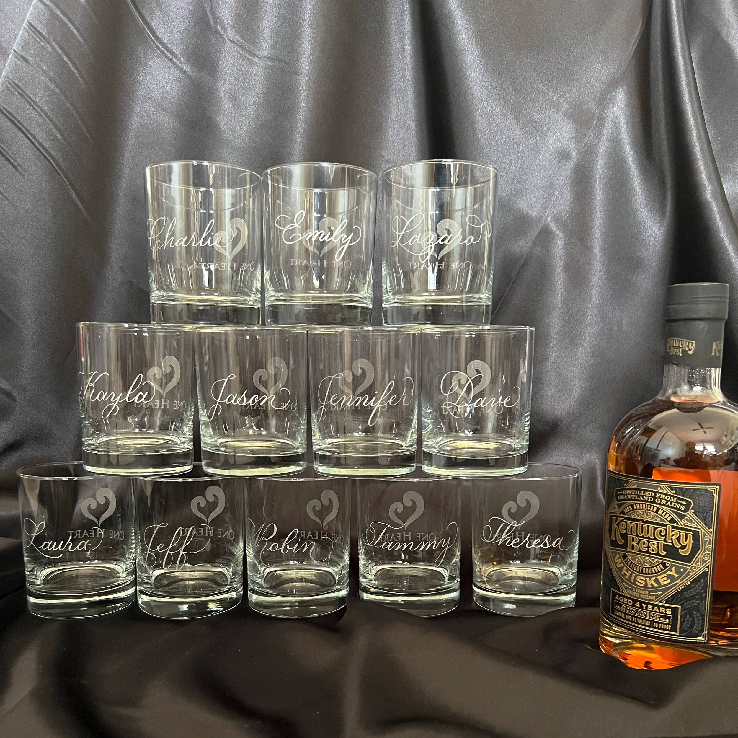Arranged whiskey glasses with names hand engraved on each glass, stacked in a pyramid shape next to a bottle of Kentucky Bourbon whiskey on a black satin background.