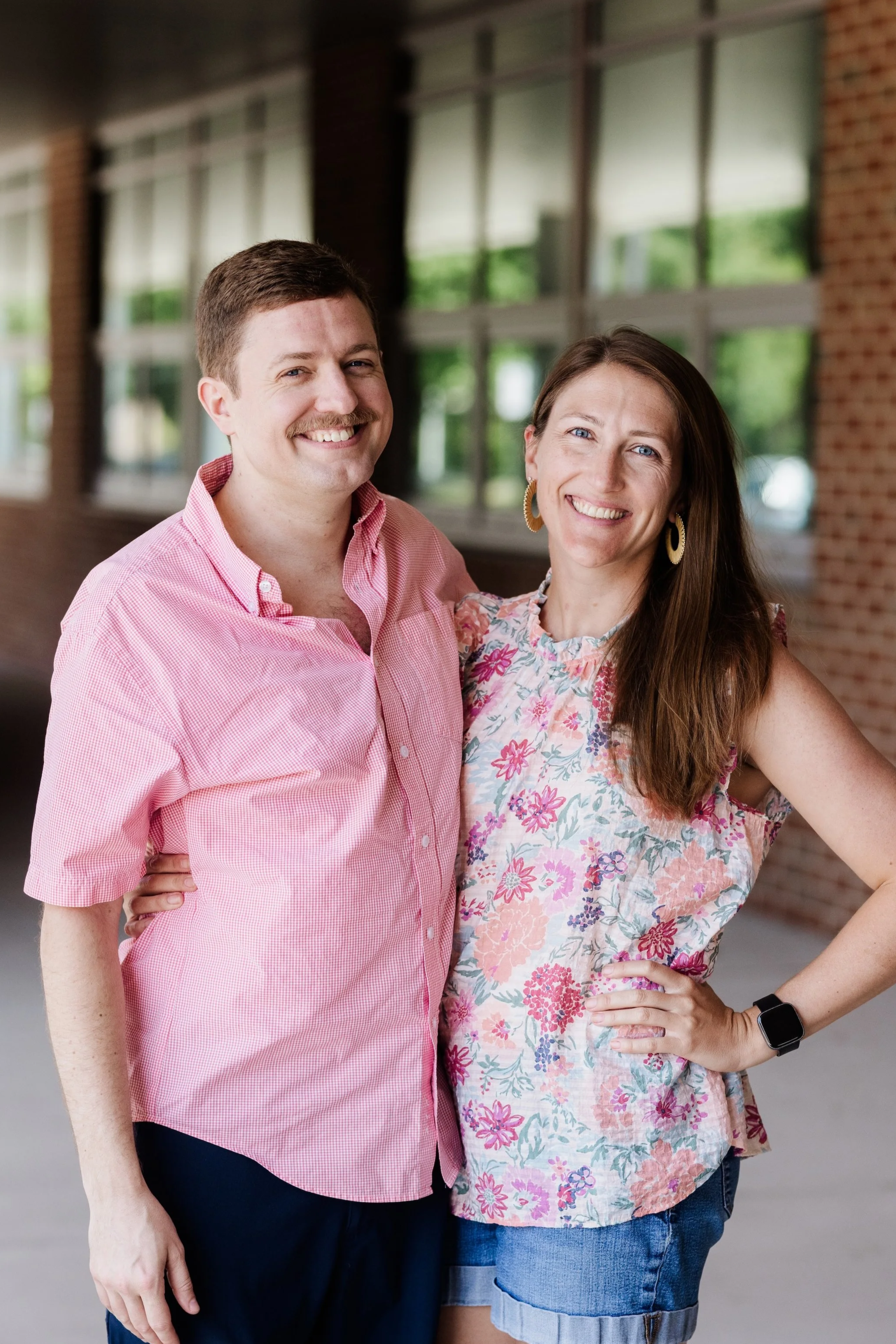 A smiling man and woman standing close together with large windows and brick wall background. The man has short brown hair and a mustache, wearing a pink checkered shirt and dark pants. The woman has long brown hair, wearing hoop earrings, a floral sleeveless top, and shorts
