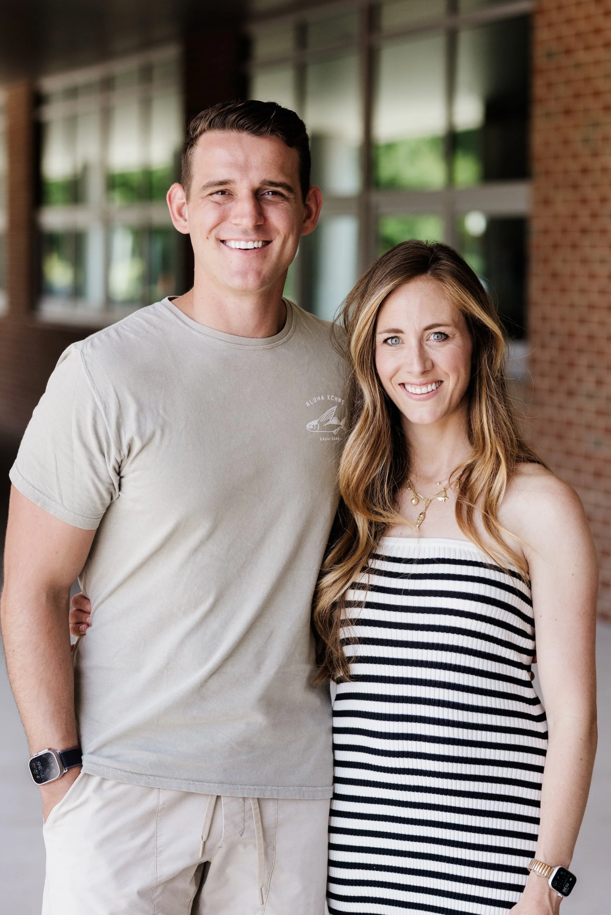 A young man and a young woman smiling and standing close together outside. The man is wearing a beige t-shirt beige shorts. The woman is wearing a stripped black and white dress. They are standing near a brick building with large windows.