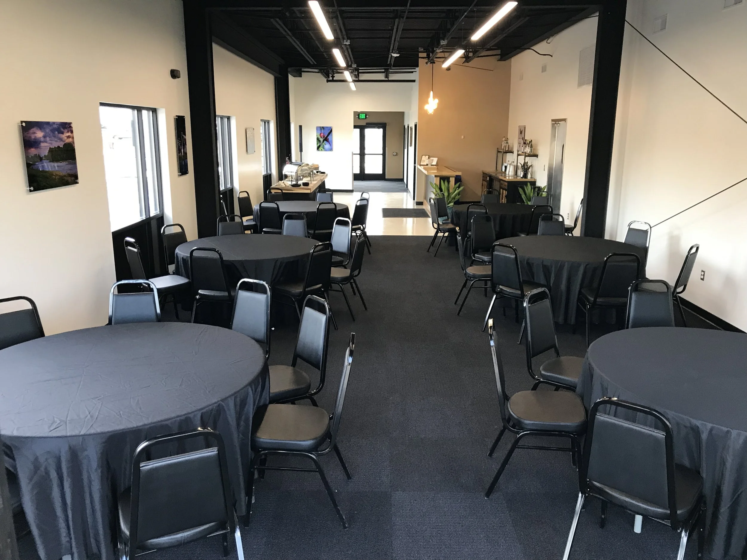A banquet hall set up with round tables covered in black tablecloths, surrounded by black chairs. The room has cream-colored walls, large windows on one side, and artwork on the walls. There is a buffet area in the back and a small bar or refreshment