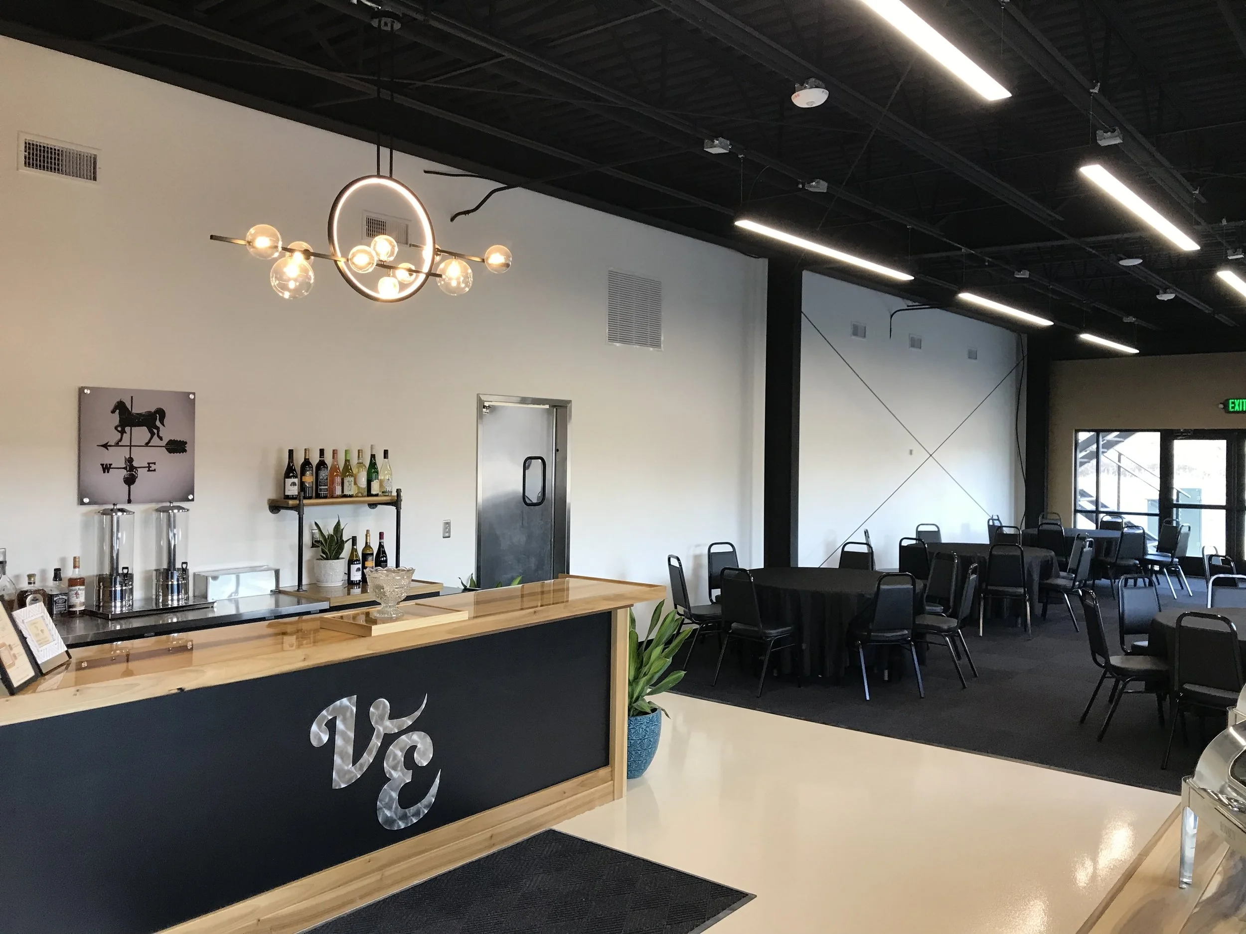Modern event space with black and wooden furniture, tables and chairs, and a bar area. The room has black ceiling with exposed ducts, white and black walls, and large windows with natural light. There is a decorative chandelier and a wall clock with