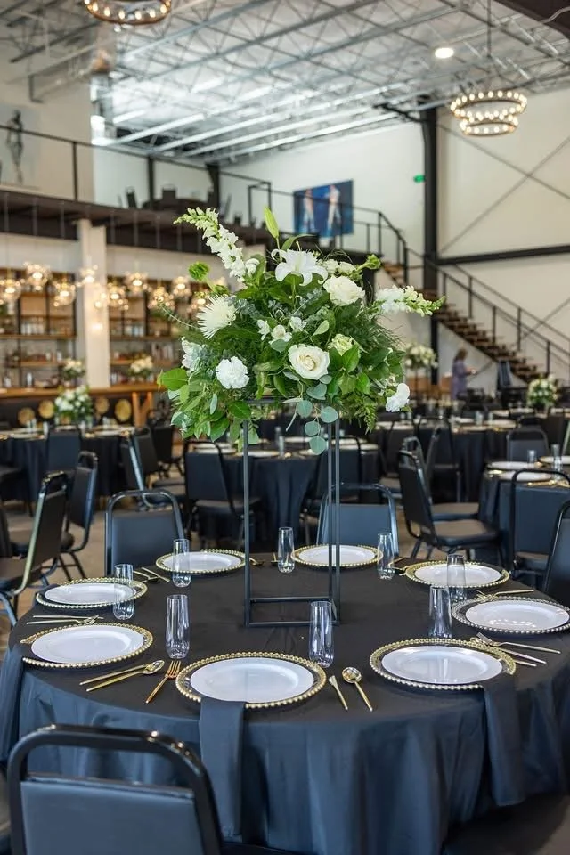 Elegant banquet table with black tablecloth, gold-rimmed plates, gold cutlery, and clear glasses, featuring a tall centerpiece with white flowers and green foliage in a spacious, industrial-style venue.