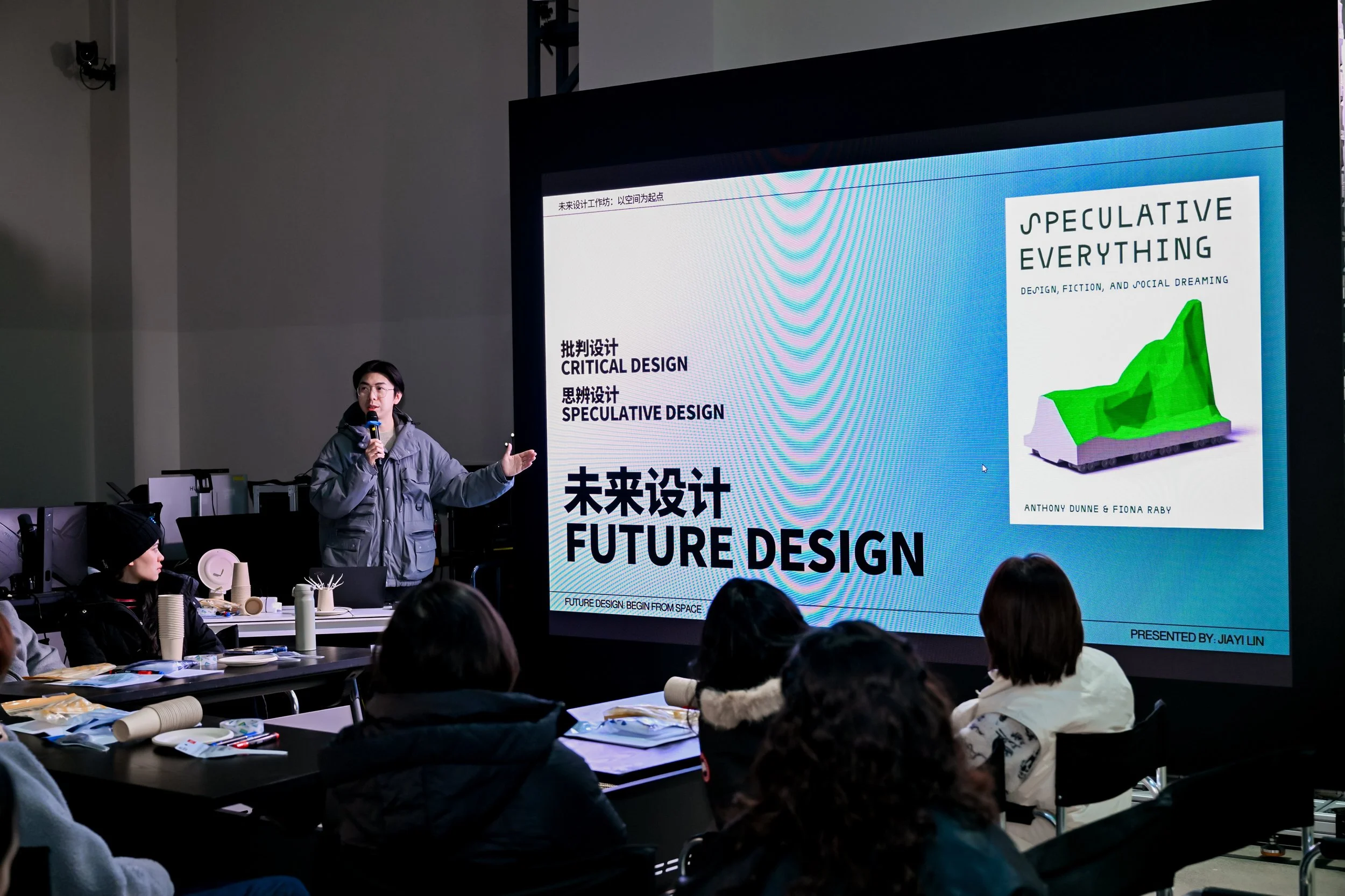 A woman is giving a presentation on future design to an audience in a conference room, with a large screen displaying slide titles including 'Critical Design' and 'Speculative Design', and a book cover titled 'Speculative Everything' by Anthony Dunne