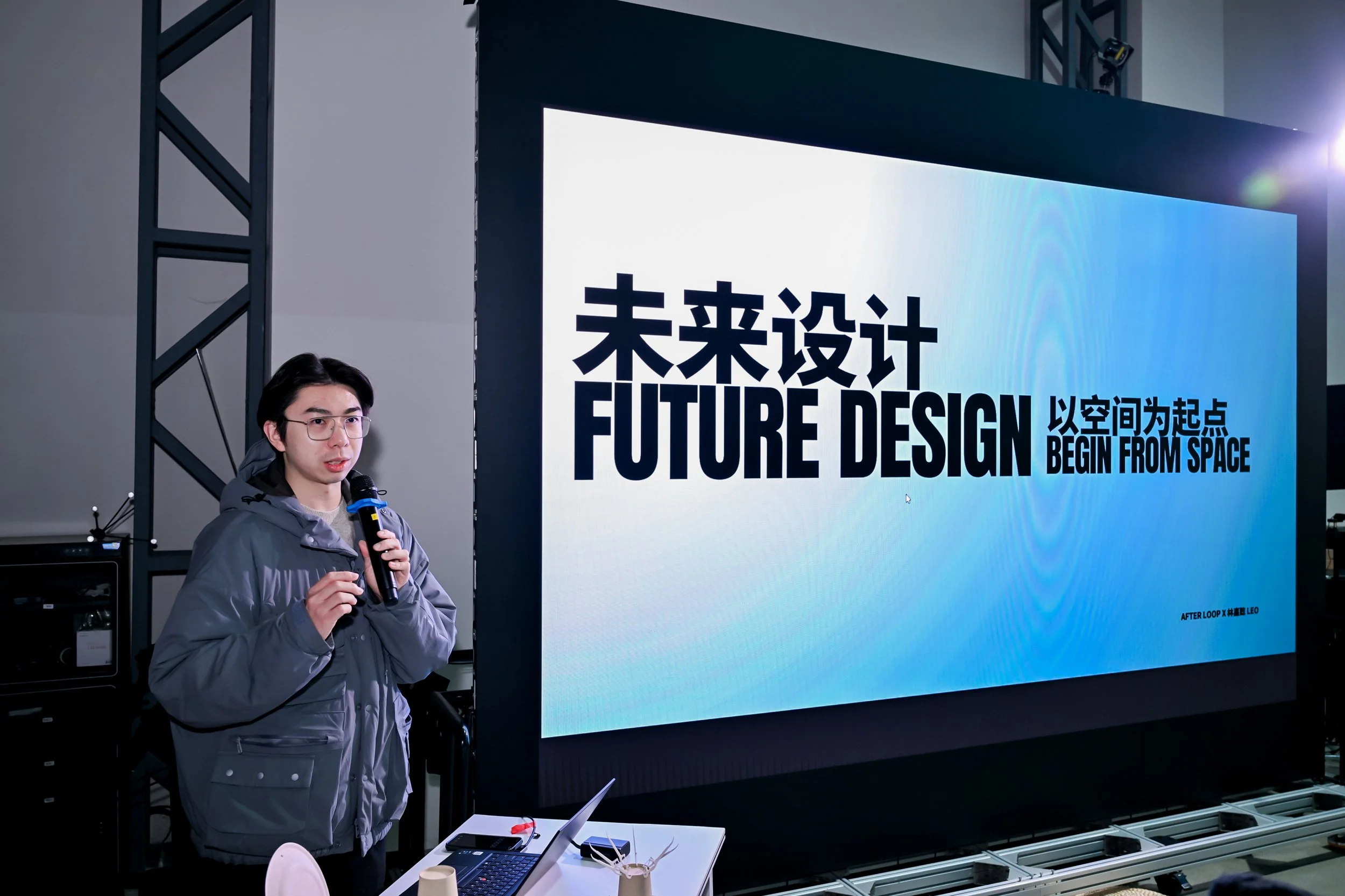A young man in a gray jacket and glasses giving a presentation with a microphone next to a large screen displaying text in Chinese and English about future design and starting from space.