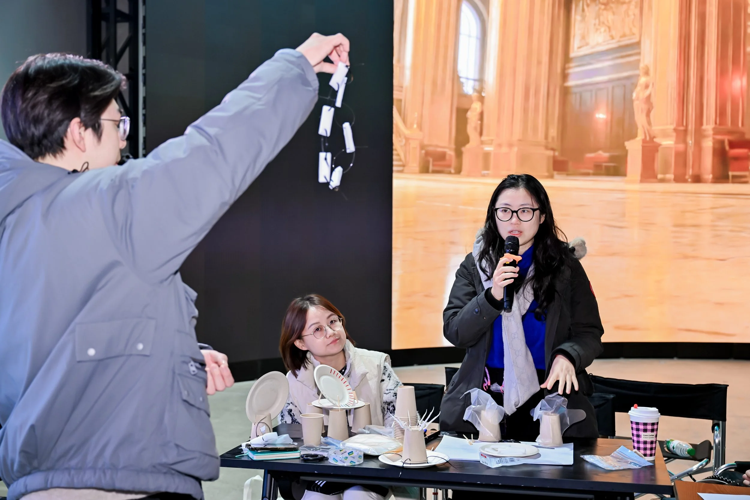 Two women are preparing for a presentation or discussion, with one woman standing and holding a microphone, and the other sitting at a table, looking up at her. The table has various paper plates, cups, and documents. A man on the left is hanging up 