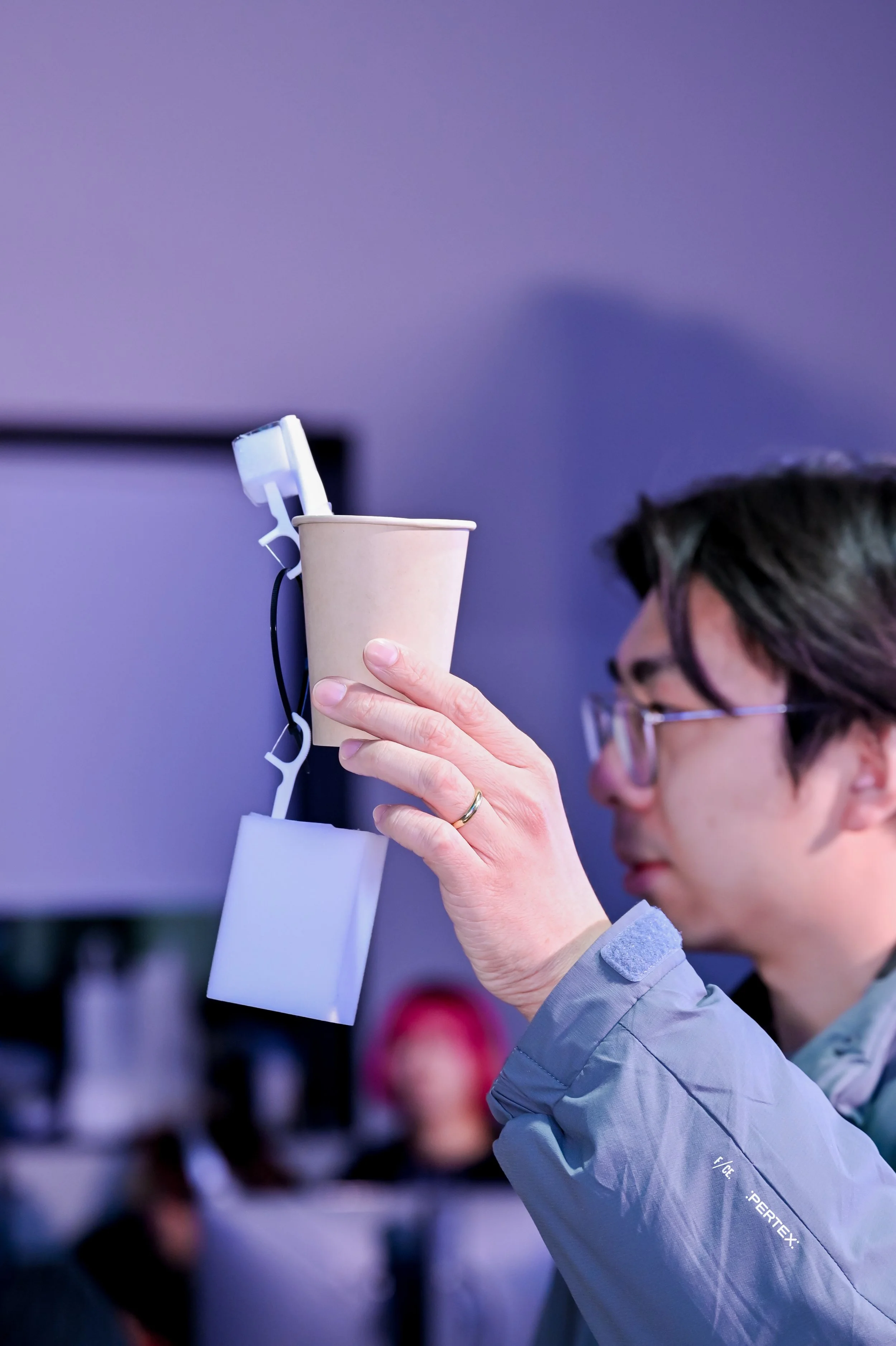 A person holding a paper cup with a smartphone and charger attached, with a blurred background of people and a blue-lit room.