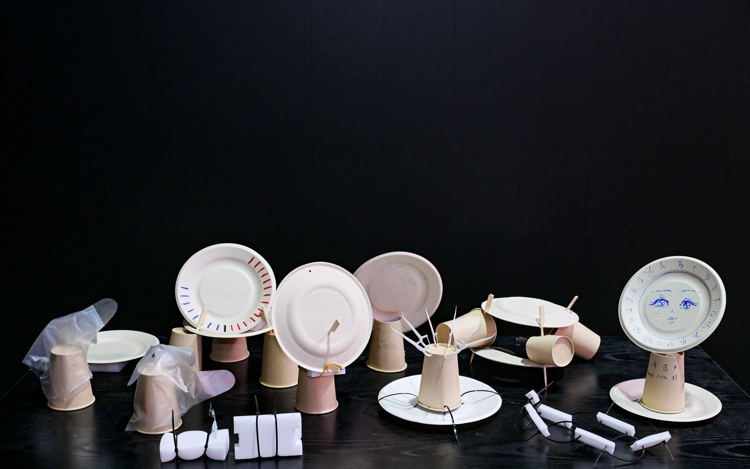 An artistic arrangement of paper plates, cups, and paper hand sculptures on a black surface against a black background. Some plates have drawings or markings, and there are string lights interwoven among the items.