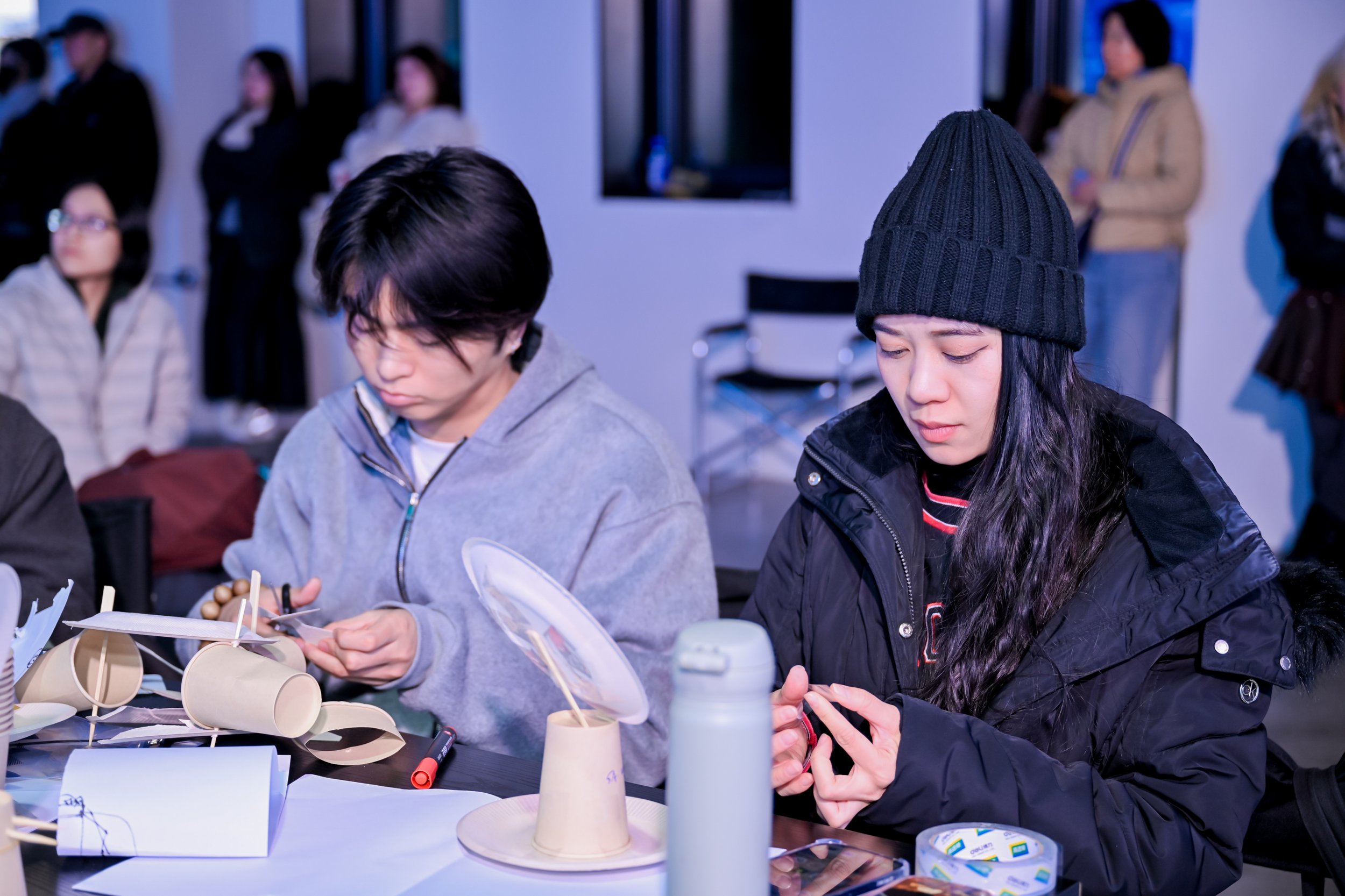 Two young women sit at a table working on a craft project involving paper cups and wooden sticks, with other people in the background.