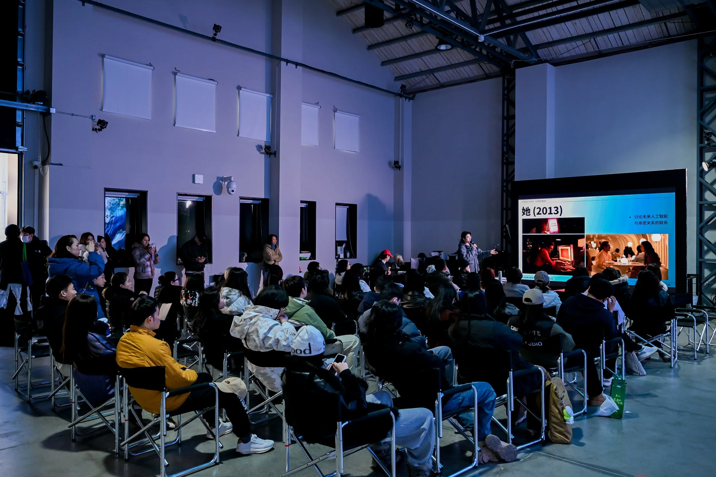A group of people sitting on chairs watching a presentation in an indoor venue with a large screen. There are a few people standing along the walls, some wearing masks. The room has high ceilings and several windows.