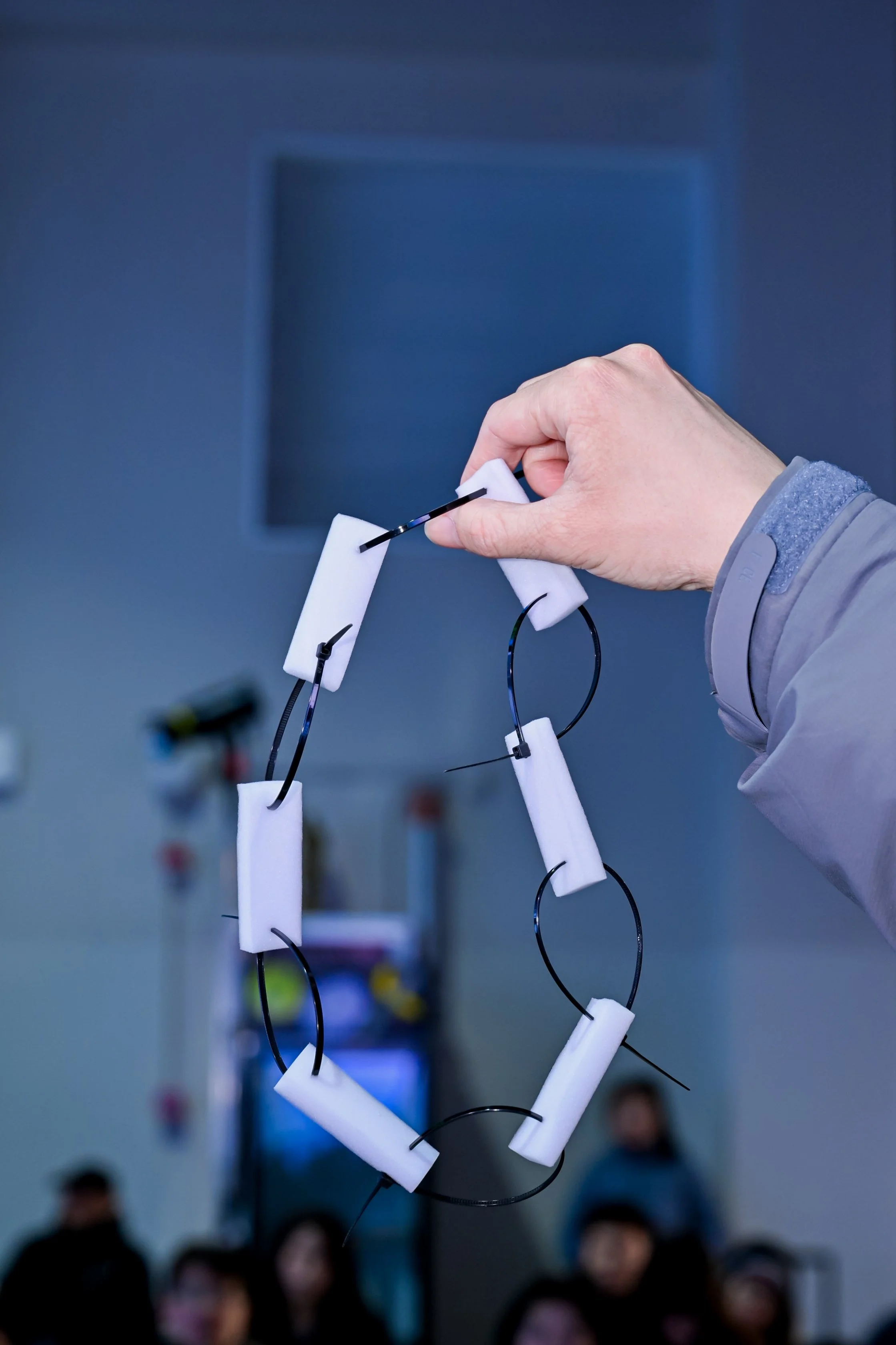 Person holding a necklace made of white foam and black wire in front of a blurred background with an audience.