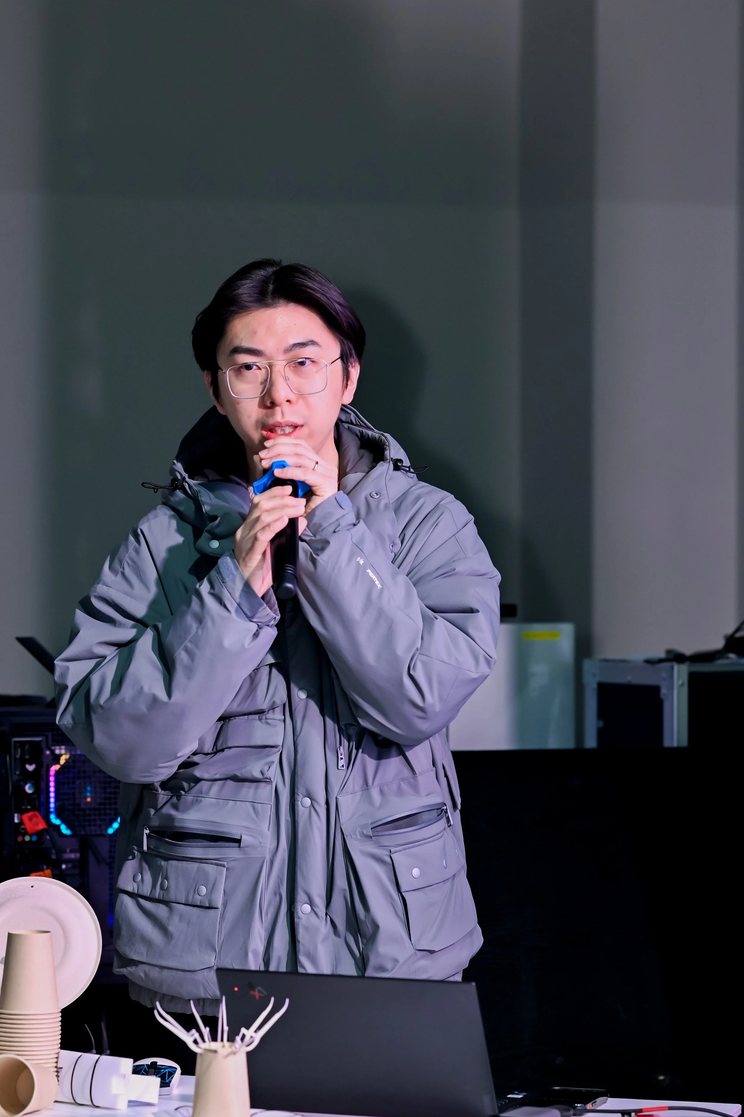 A young man with glasses speaking into a microphone in a room with technology and cups on a table.