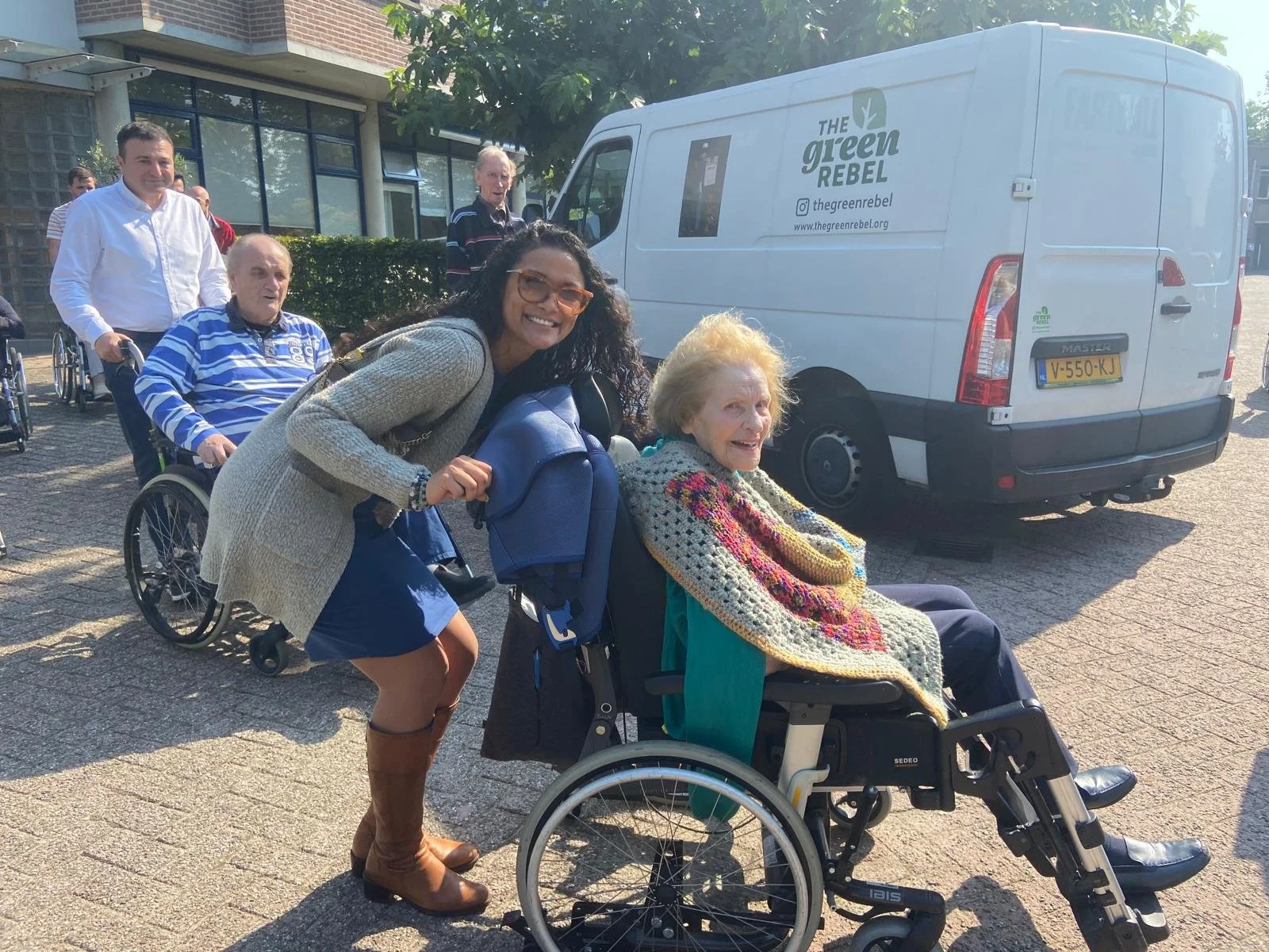 Een vrijwilliger en dame in rolstoel kijken lachend in de camera.