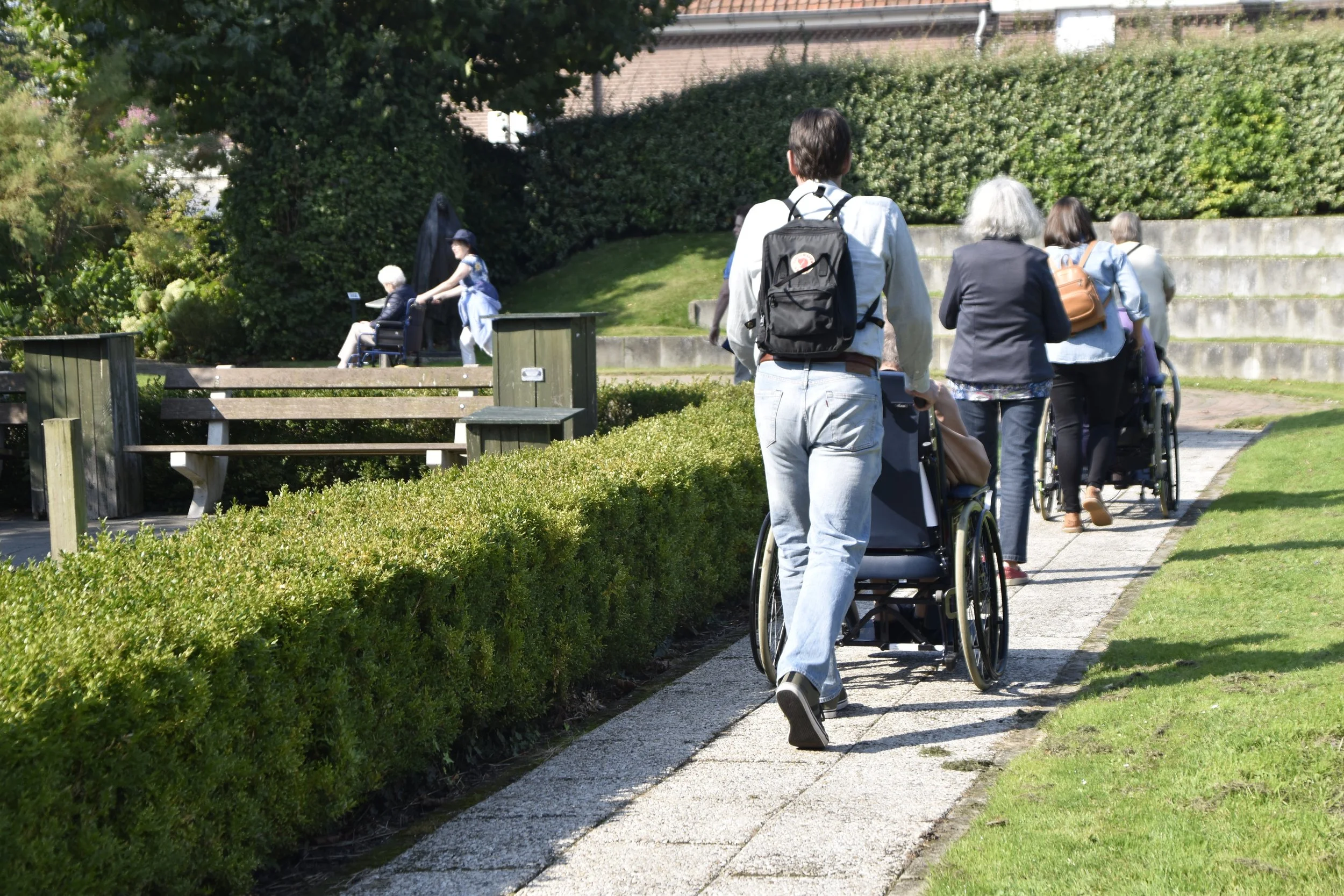 Vrijwilligers rijden ouderen in rolstoel door een groen park.