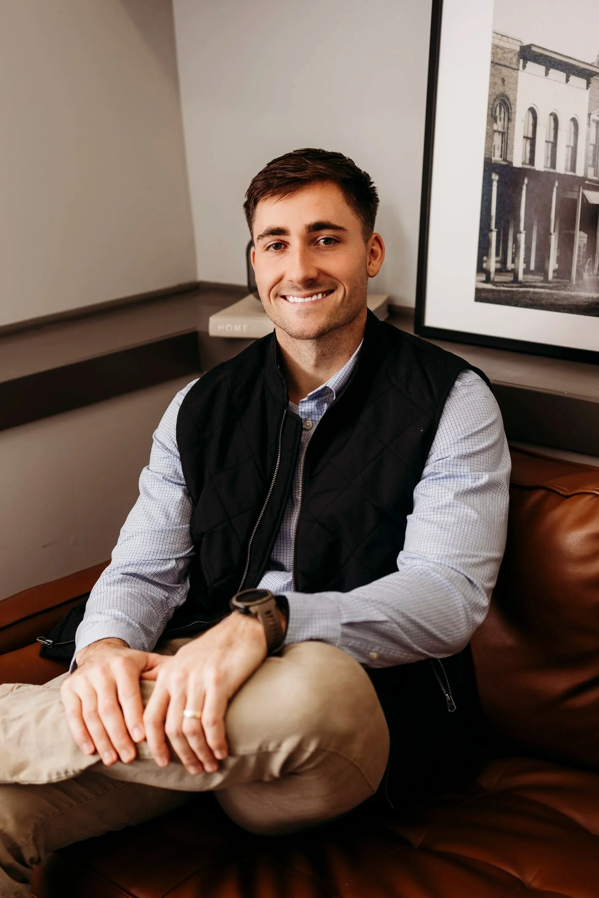 Portrait of a smiling young man with crossed arms, wearing a black vest over a light checkered shirt, standing against a plain gray background.