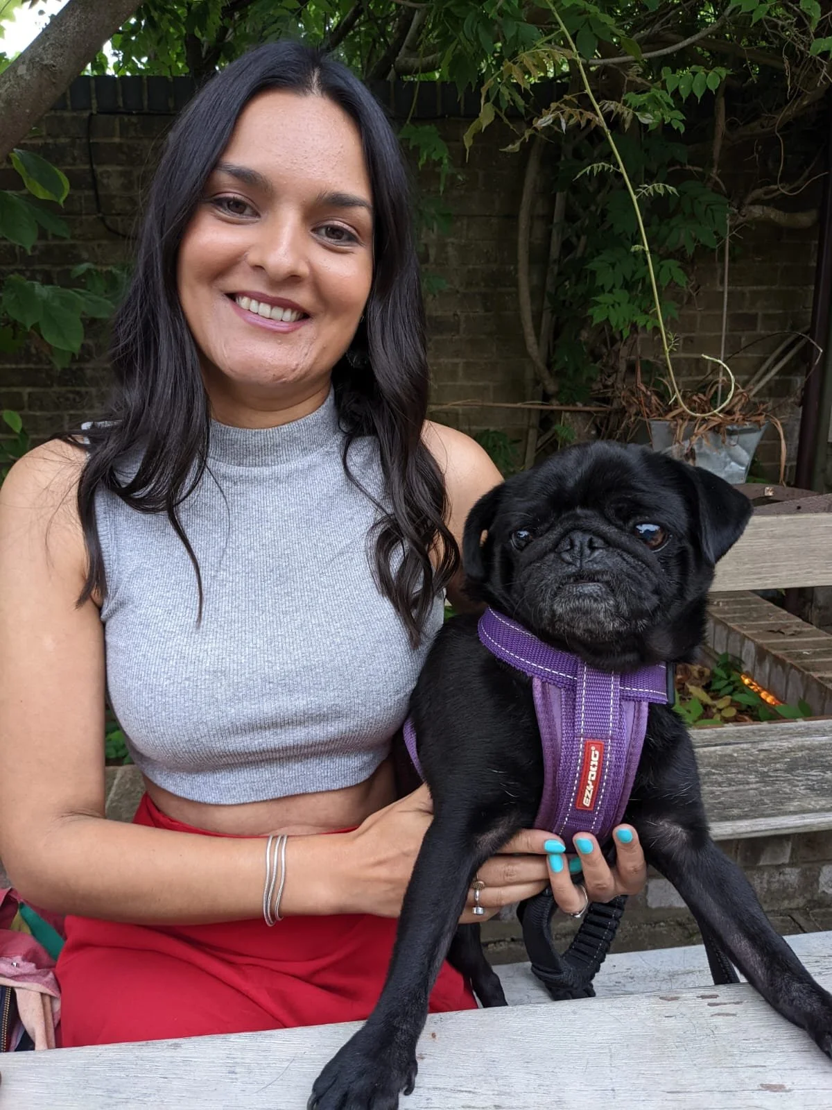 Asha, a Brown-skinned woman, smiles at the camera holding her black pug. She's sitting at a wooden bench with a wall and trees behind her.