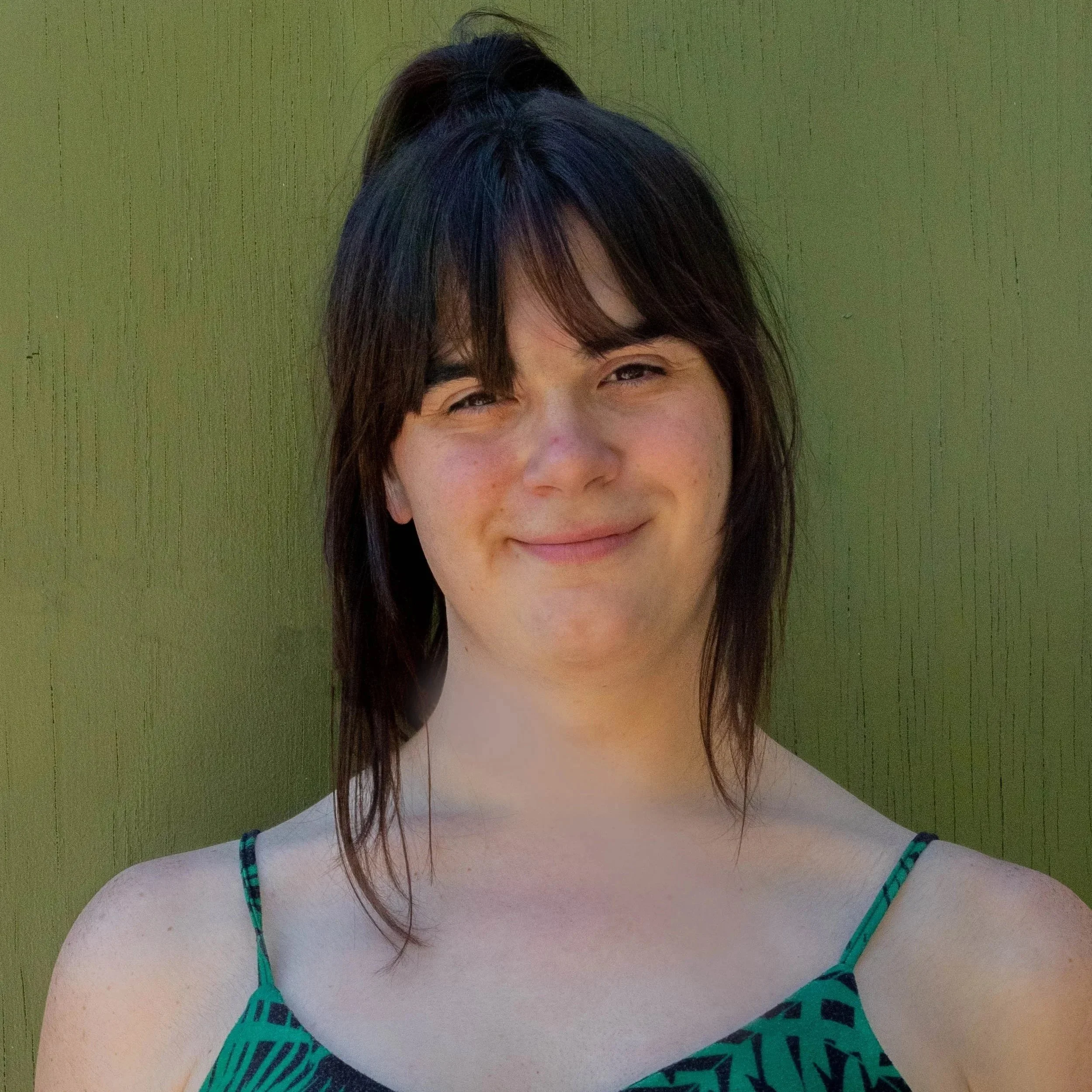 Young woman with dark brown hair and bangs, smiling, wearing a green and black patterned sleeveless top, stands against a green background.