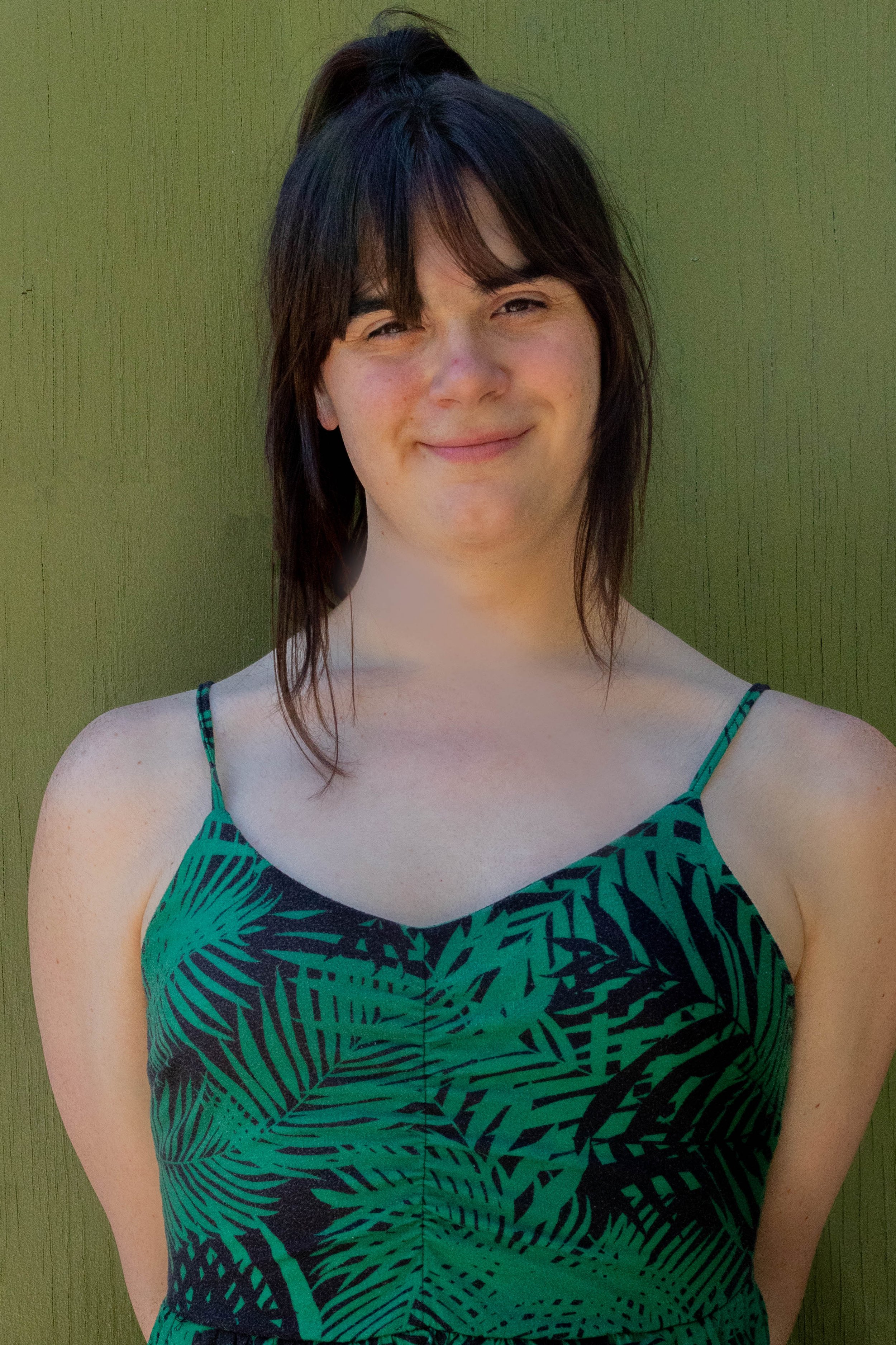 A young woman with dark brown, shoulder-length hair and a friendly smile, wearing a green and black patterned spaghetti strap dress, standing in front of a green wooden wall.