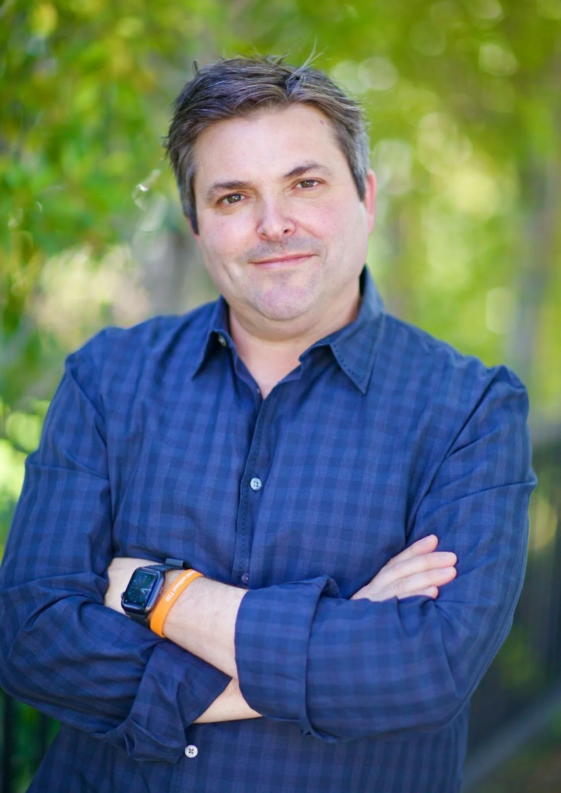 A man with dark hair in a blue button-up shirt standing outdoors with his arms crossed, smiling slightly, with a background of green trees and foliage.
