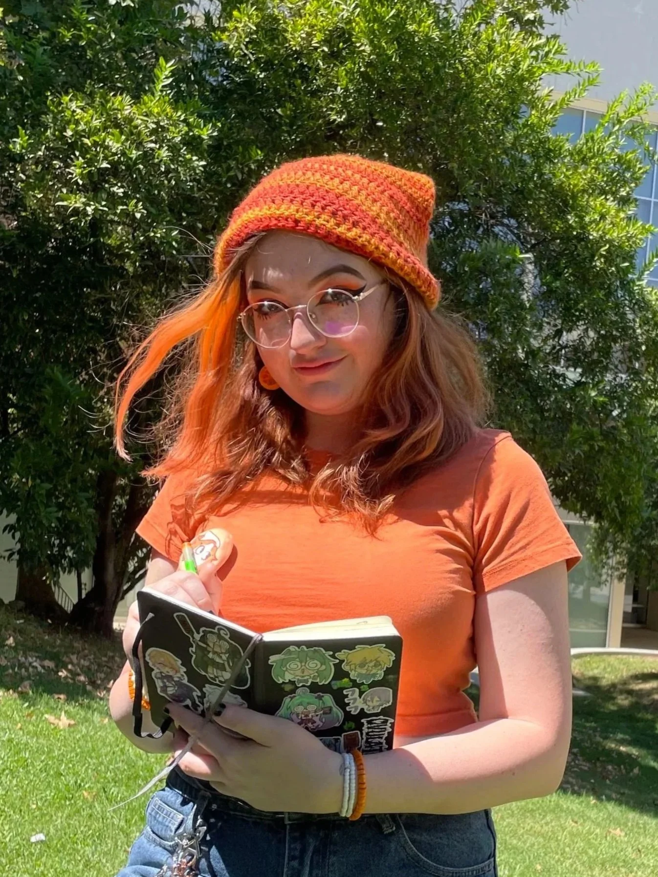 A young woman with red and orange hair wearing a colorful crochet beanie, glasses with round frames, and an orange t-shirt, standing outdoors in front of green trees and a building, holding a black notebook with stickers and writing in it.