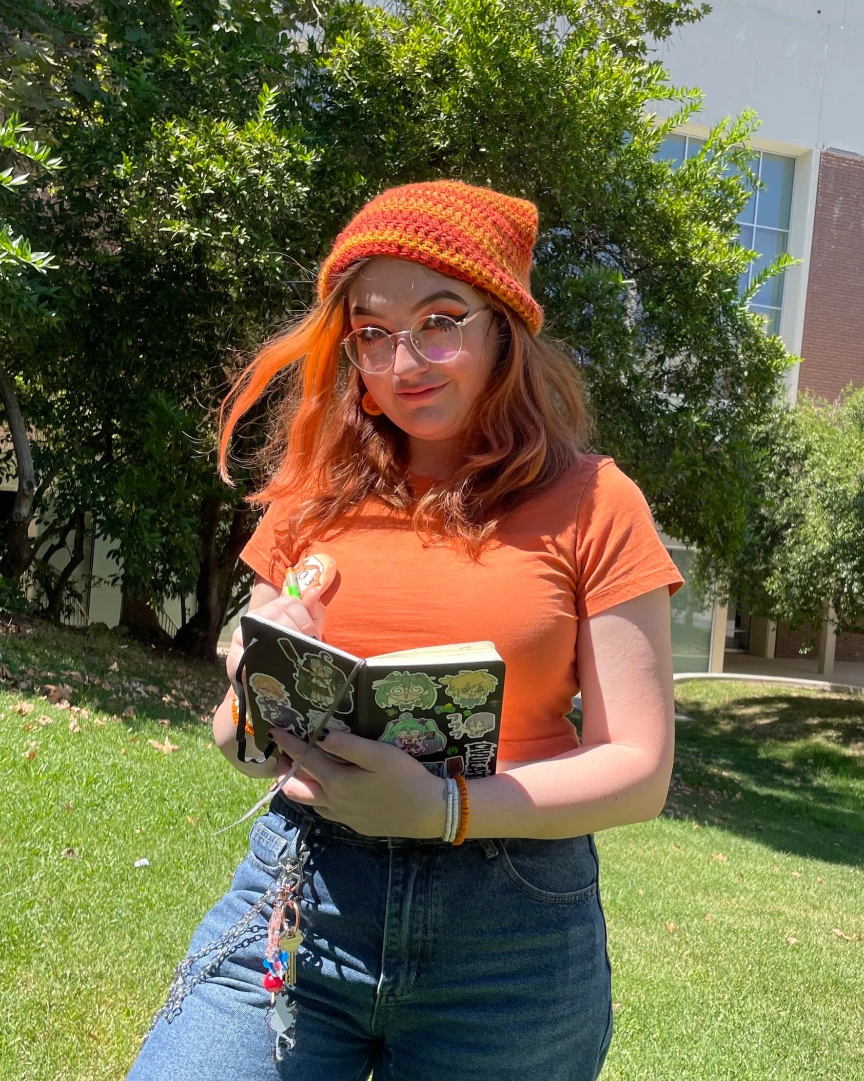 A young woman with orange hair, wearing glasses, a colorful knit beanie, an orange T-shirt, and high-waisted jeans, is standing outdoors on a grassy area with trees and a building in the background. She is holding a small notebook with stickers and a green pen.