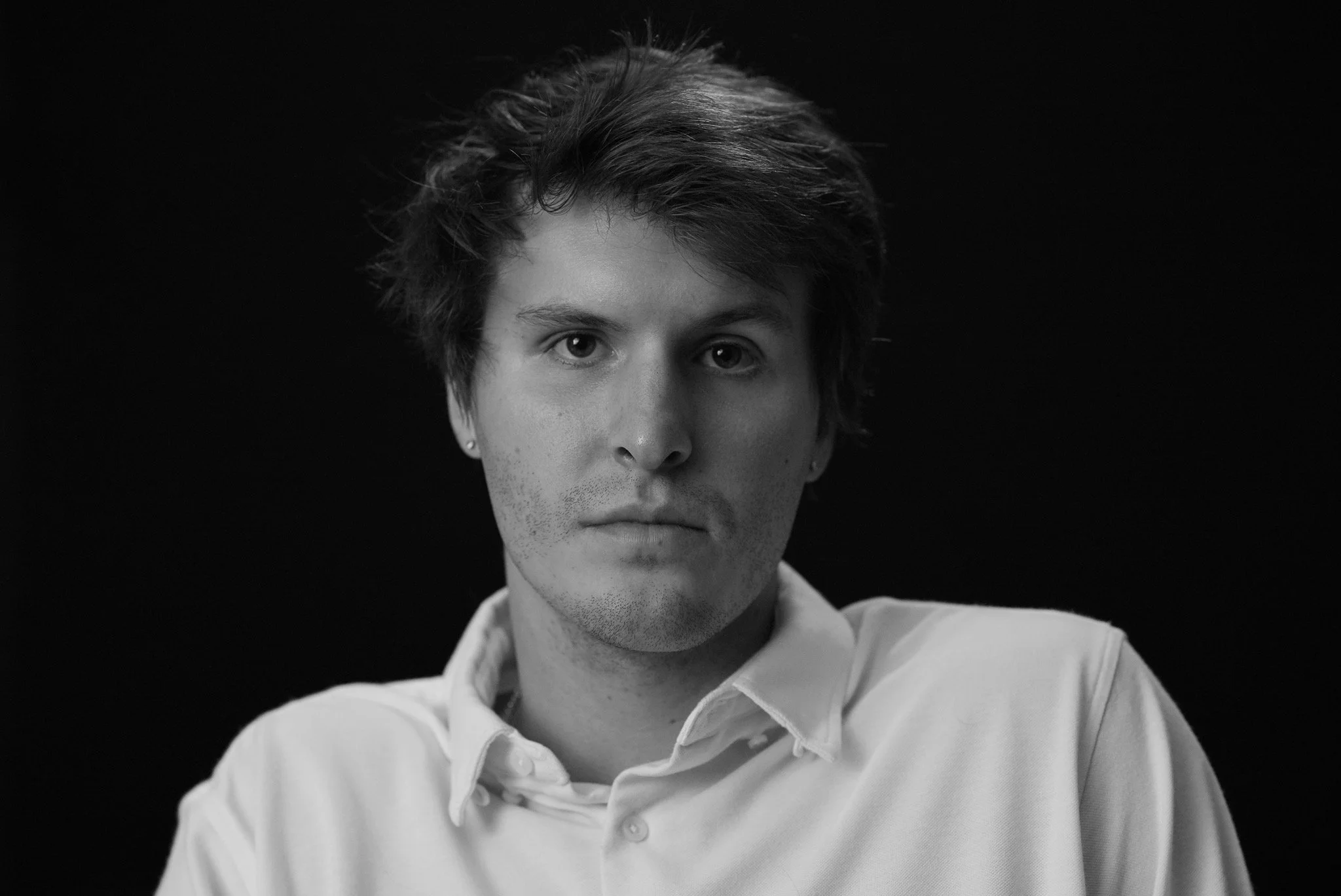 Black and white portrait of a young man with dark, tousled hair, wearing a white collared shirt, looking directly at the camera against a black background.